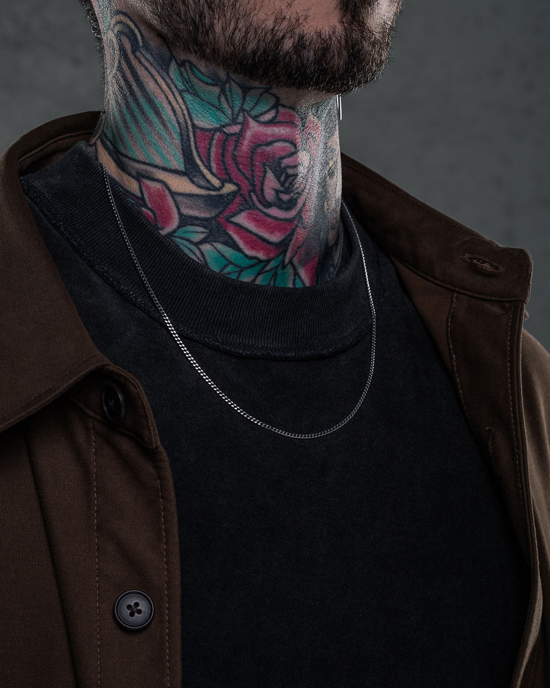 Wearing a brown jacket and black shirt, a person with a colorful rose-and-leaf neck tattoo accessorizes with a 2.3mm Curb Chain necklace. The photo focuses on the neck, clothing, and necklace, with the face partially visible.