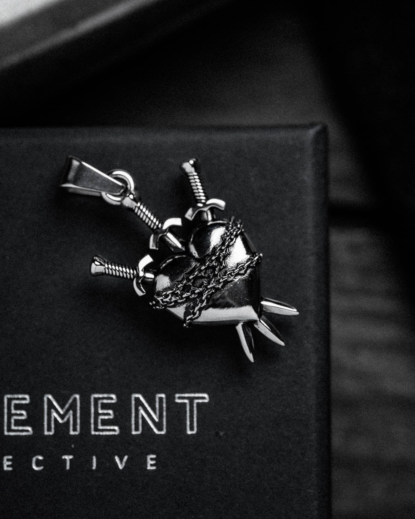 The "3 of Swords" Pendant is a metallic heart pierced by three swords and wrapped in chains, displayed on a black surface with partial white text.