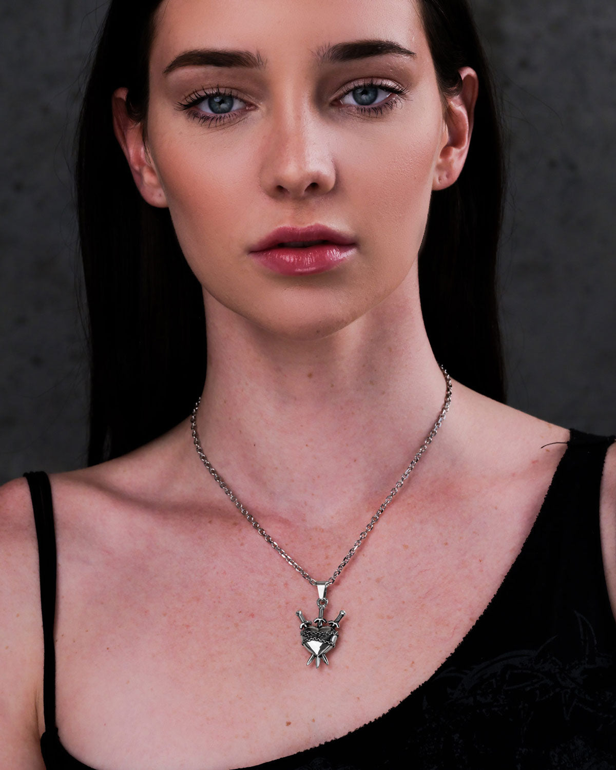 A woman with long dark hair wears a black top and the "3 of Swords" Pendant—a silver necklace with a heart-shaped pendant and two crossed swords—standing against a plain, dark background.