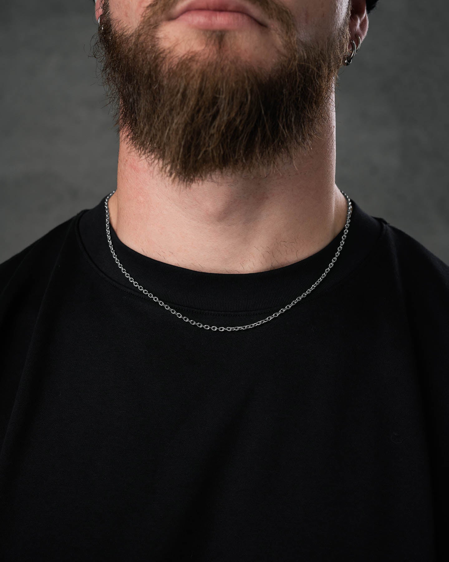 A close-up of a man in a black shirt wearing the 3mm Cable Chain, a thin, non-tarnish stainless steel necklace resting just above his collarbone. He has a full brown beard and stands against a plain dark gray background, face partially out of frame.
