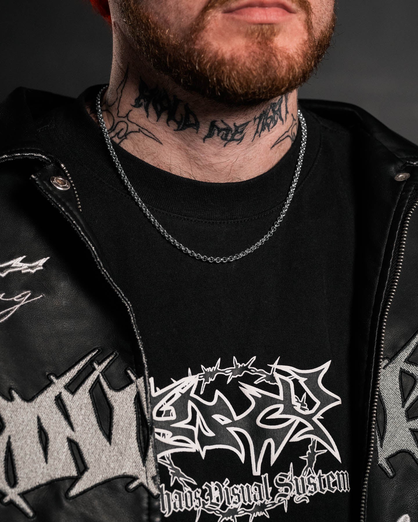 A bearded person with neck tattoos wears a 4mm Rolo Chain, black graphic tee, and black leather jacket with white graffiti text. Image is cropped to show only the lower face and upper torso.