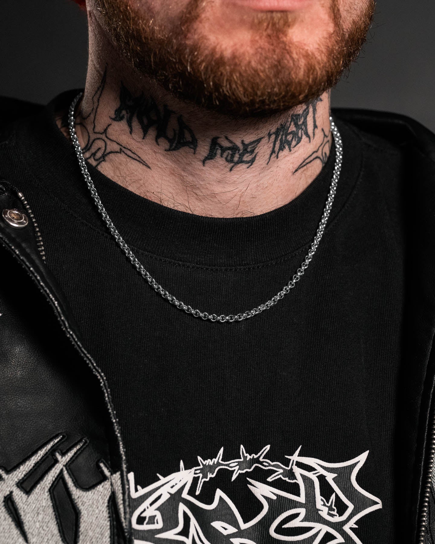 A close-up of a bearded man’s lower face and neck shows a tattoo and a silver 4mm Rolo Chain necklace. He wears a black t-shirt with a bold white graphic under a black leather jacket.