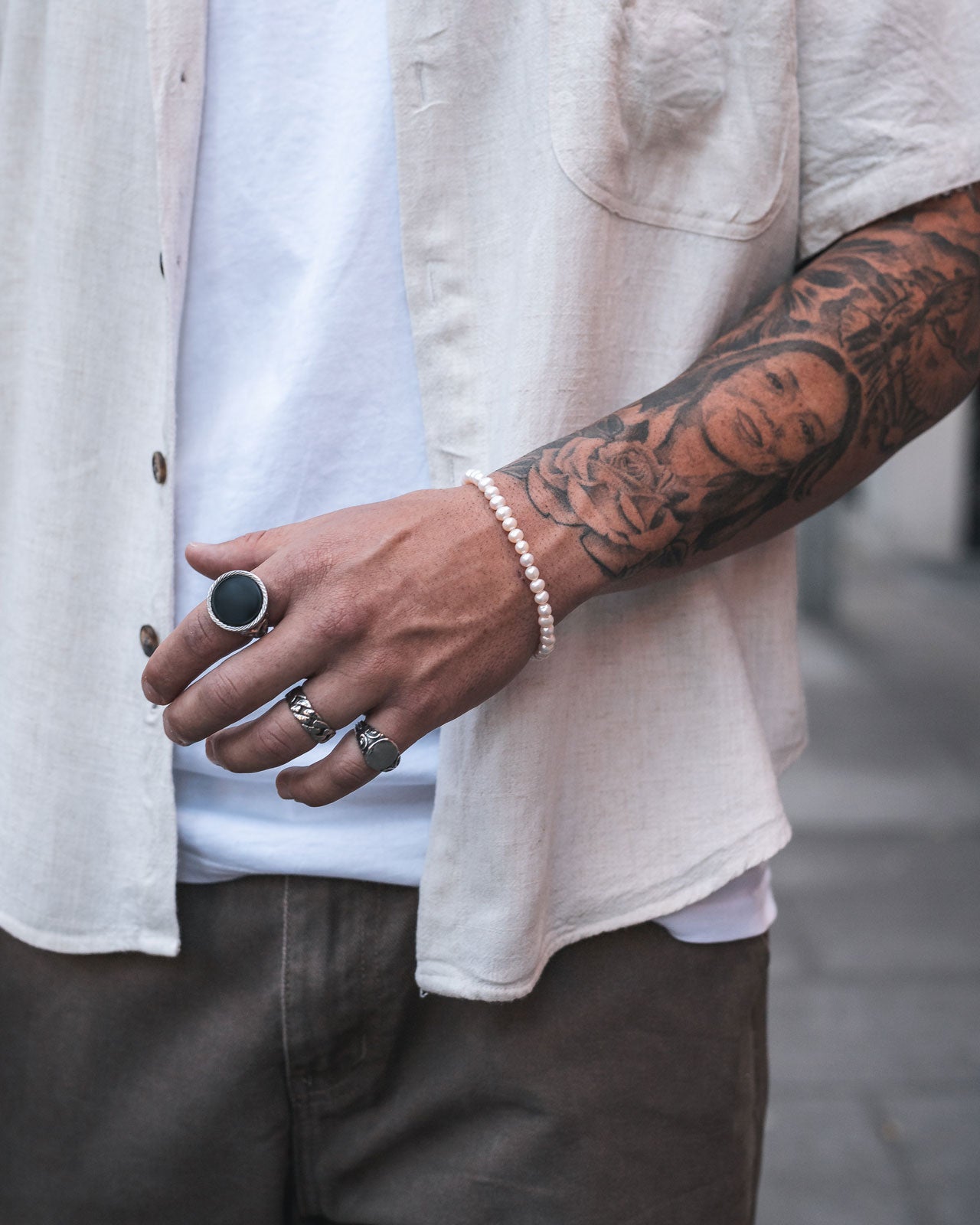 A person in a white shirt and brown pants displays a tattooed arm with rings and a 5mm Freshwater Pearl Bracelet. The rose and face tattoos are visible as the relaxed hand extends to showcase the elegant accessory.