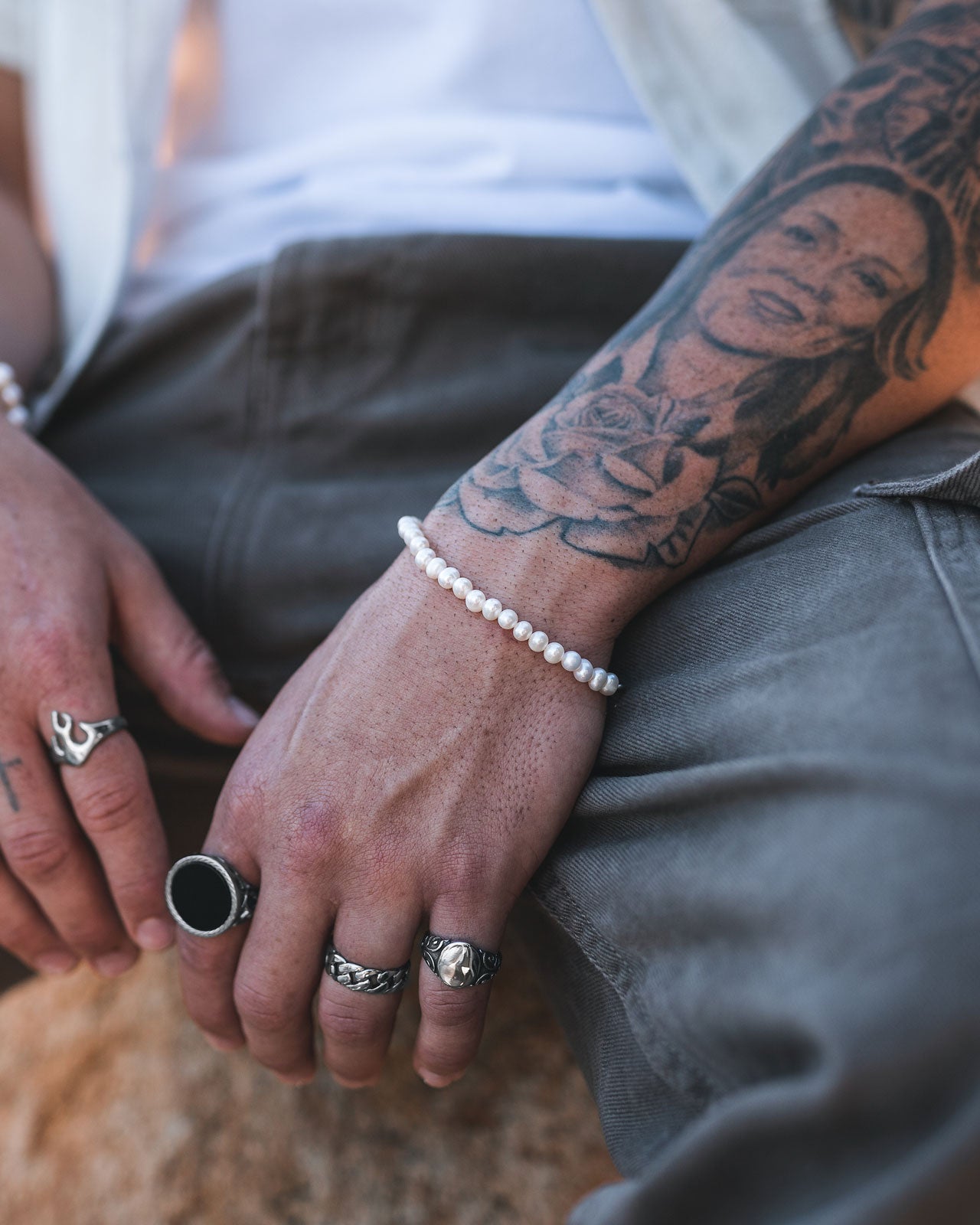 A close-up of a person’s hands and forearm shows multiple silver rings, a 5mm Freshwater Pearl Bracelet, and a detailed tattoo of a woman’s face and rose. The person is wearing a white shirt and gray pants.