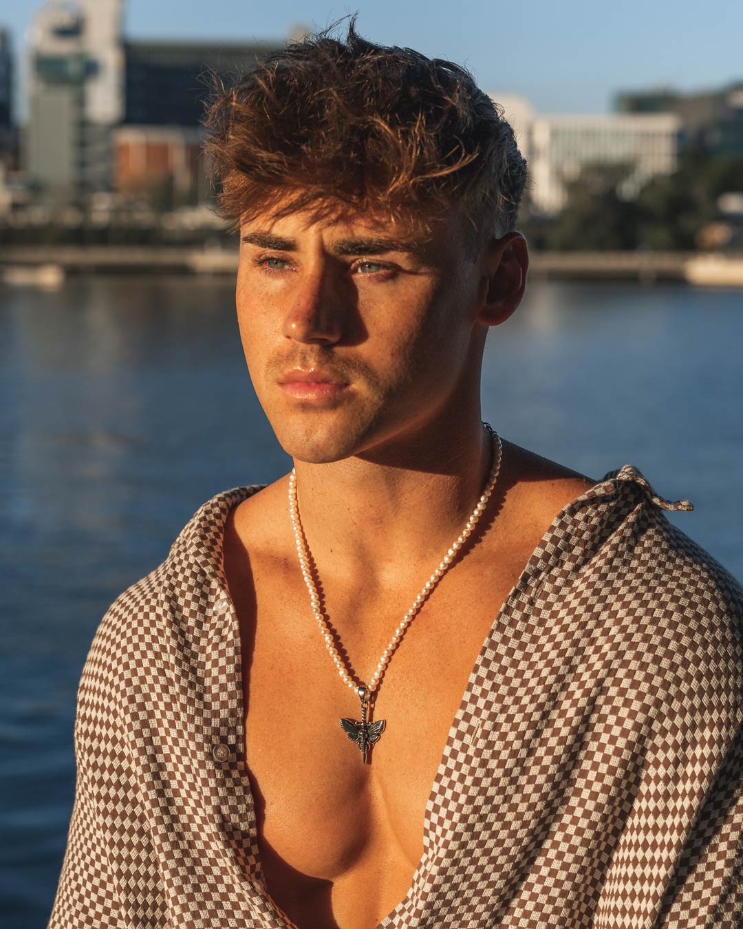 A young man with tousled brown hair stands by the water, sunlight on his face, wearing an open checkered shirt and a Freshwater Pearl Necklace With Custom Pendant Choice. City buildings can be seen in the background as he looks thoughtfully into the distance.