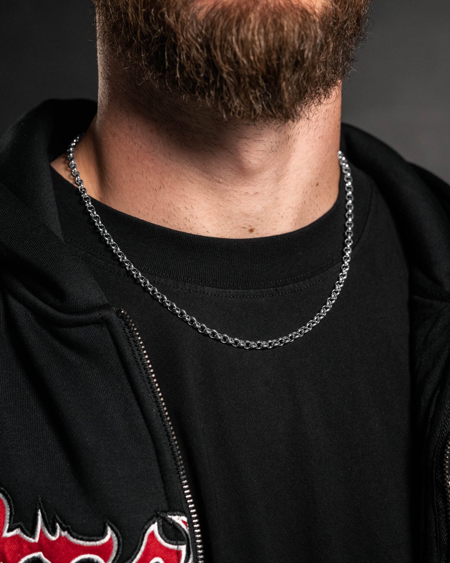 A close-up of a person wearing the 5mm Rolo Chain necklace, along with a black t-shirt and a black zip-up jacket featuring a red and white embroidered design. The lower face and neck with a brown beard are visible.