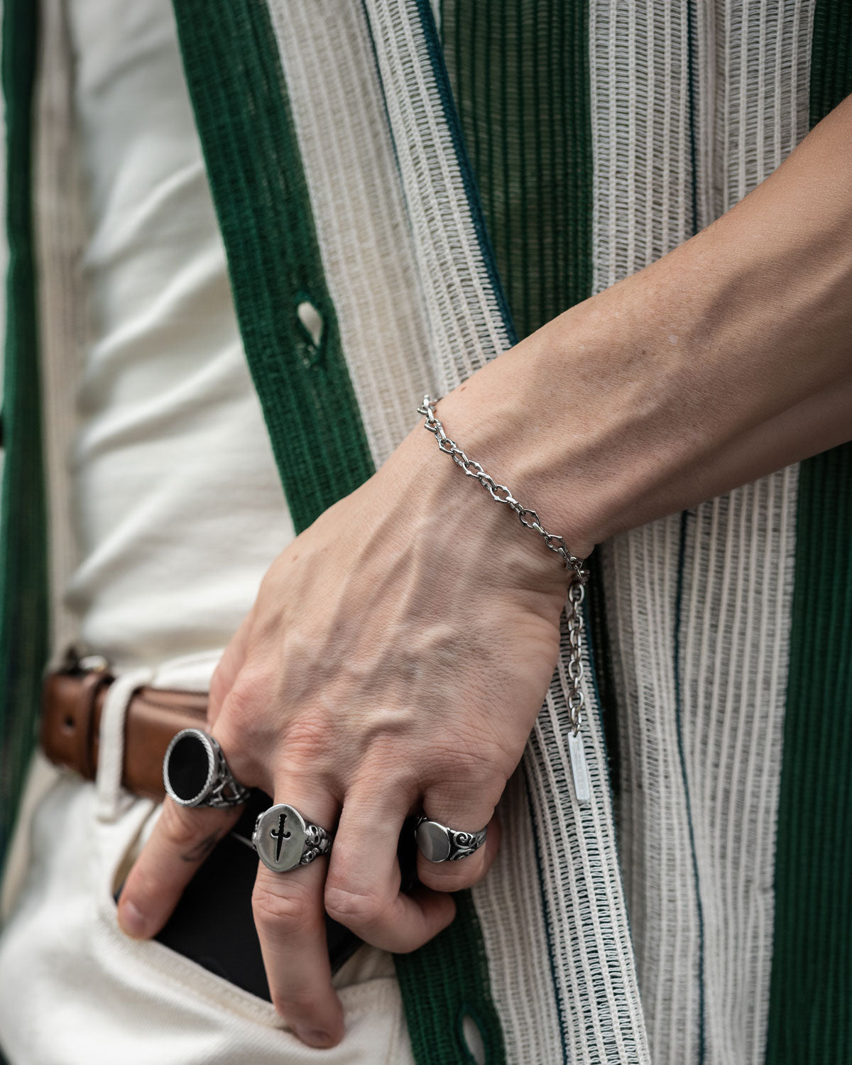 A close-up of a hand wearing silver rings and "The Cathedral" 6mm Spiked Bracelet, holding a phone. The person is dressed in a green and white vertically striped shirt with a brown belt.