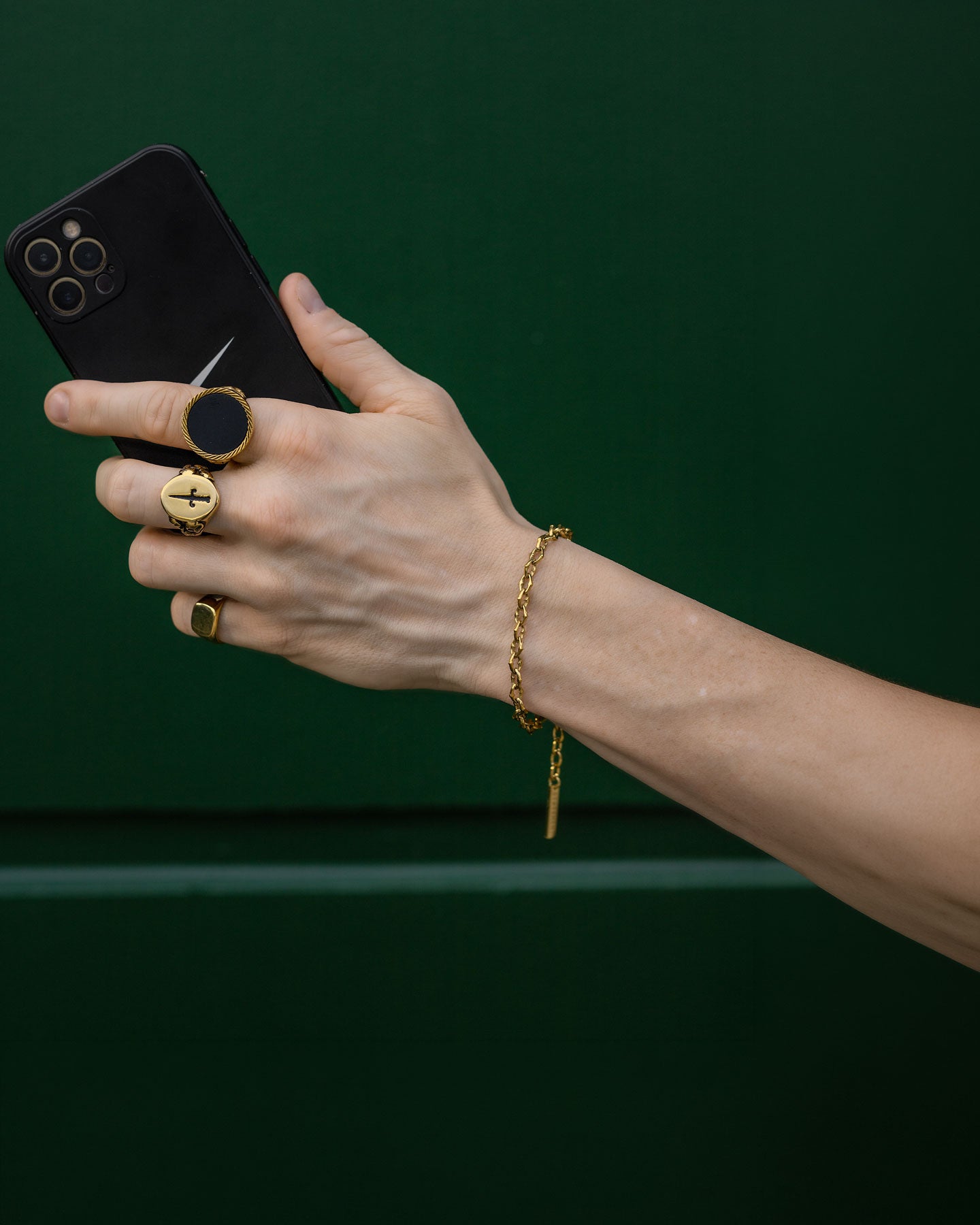 A hand wearing "The Cathedral" 6mm Spiked Bracelet (Gold) and gold rings holds a black smartphone against a solid dark green background.