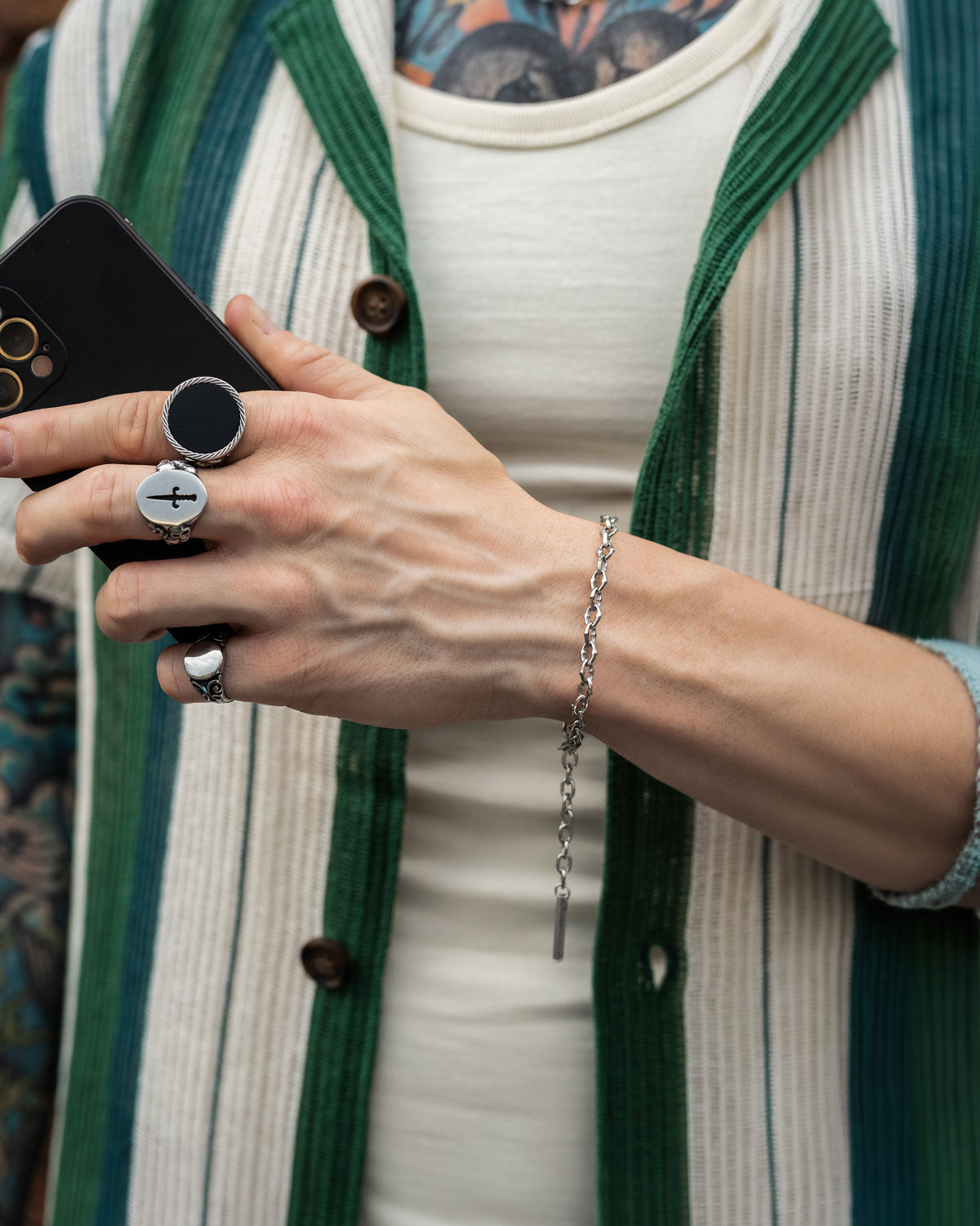 A person wearing a cream and green striped shirt holds a black smartphone. Their hand displays silver rings, including one with a cross, and "The Cathedral" 6mm Spiked Bracelet. A colorful tattoo is visible on their chest and arm.