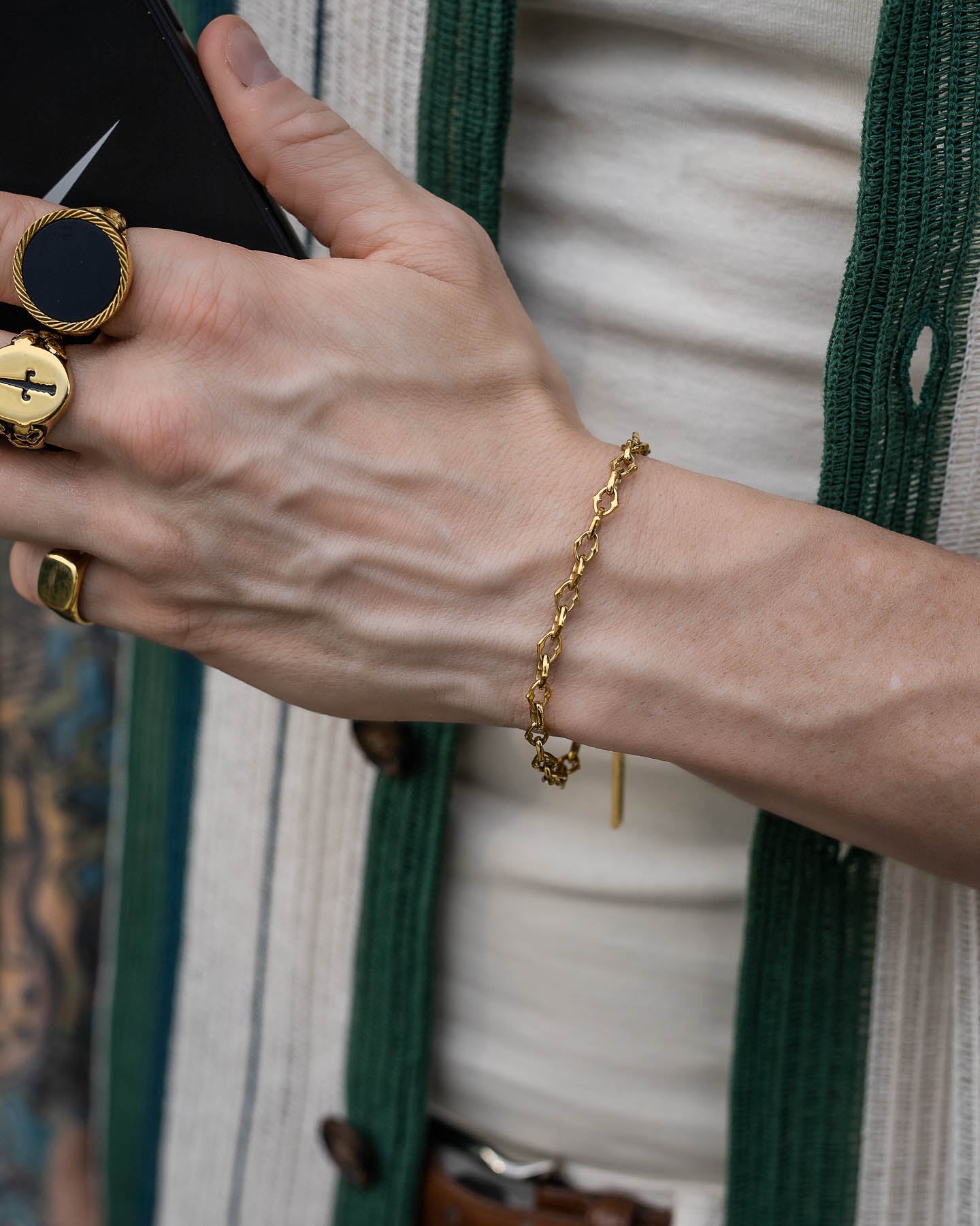 A person wearing "The Cathedral" 6mm Spiked Bracelet (Gold) and gold rings holds a black smartphone. They are dressed in a cream shirt and a green and white cardigan.