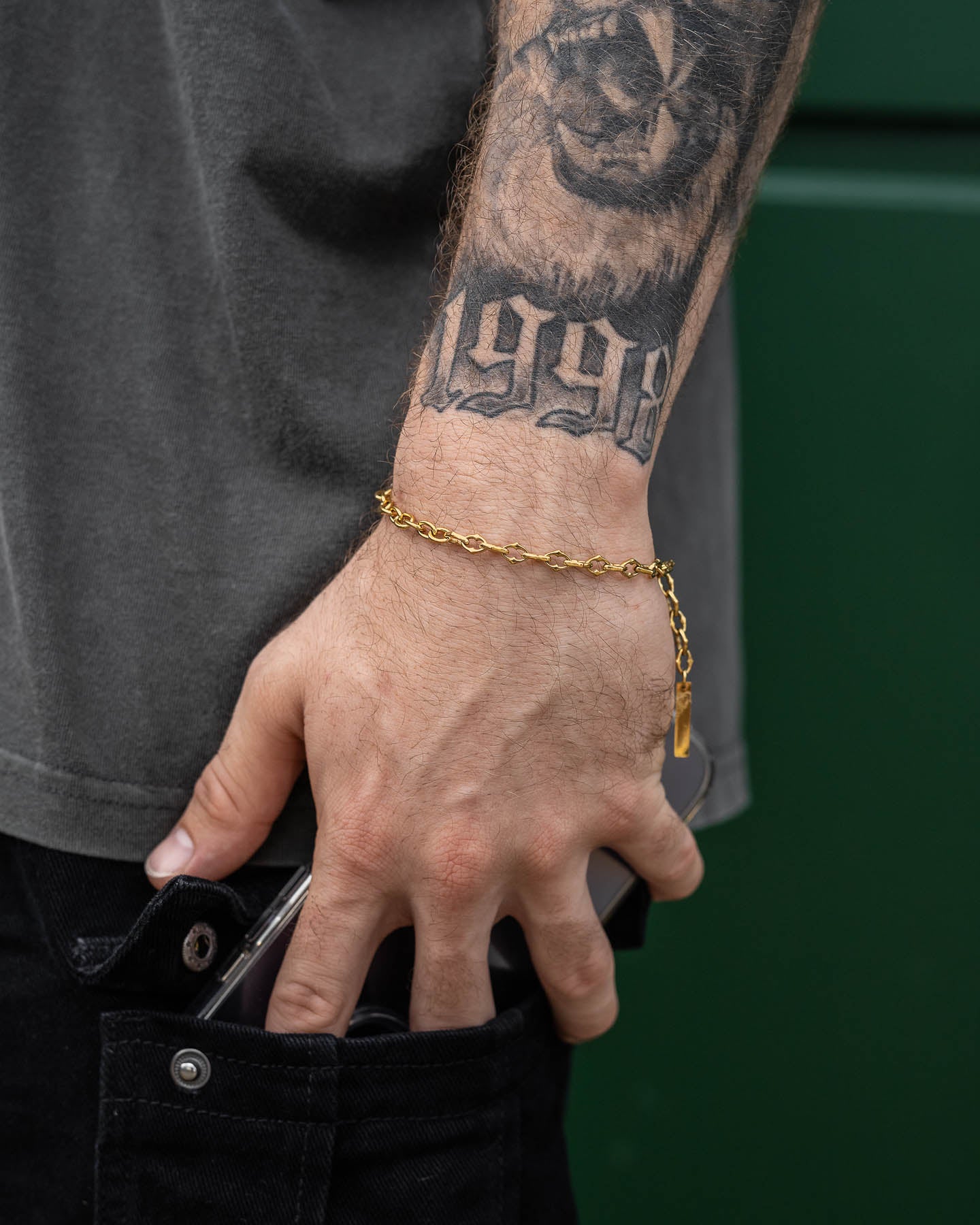 A person with a 1998 and tiger face forearm tattoo wears "The Cathedral" 6mm Spiked Bracelet (Gold) while slipping a smartphone into the front pocket of black jeans, set against a green background.