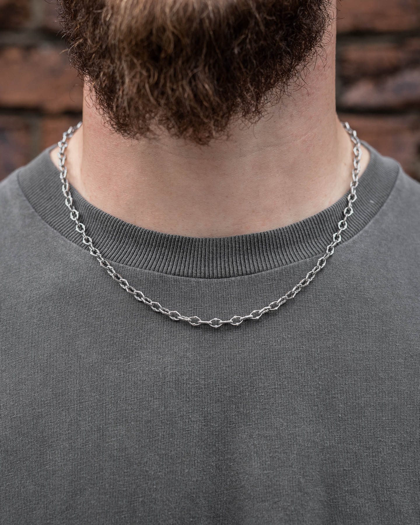 A close-up shows a person wearing "The Cathedral" 6mm Spiked Chain and a gray crew-neck shirt. The lower face and neck are visible, with a blurred reddish-brown brick wall in the background.