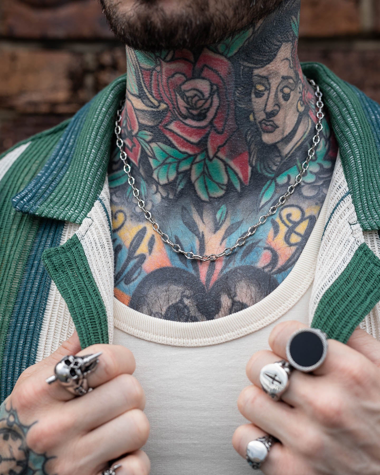 A close-up of a tattooed neck and chest with colorful rose and woman's face tattoos. The person wears silver rings, a white shirt, green striped jacket, and "The Cathedral" 6mm Spiked Chain necklace.
