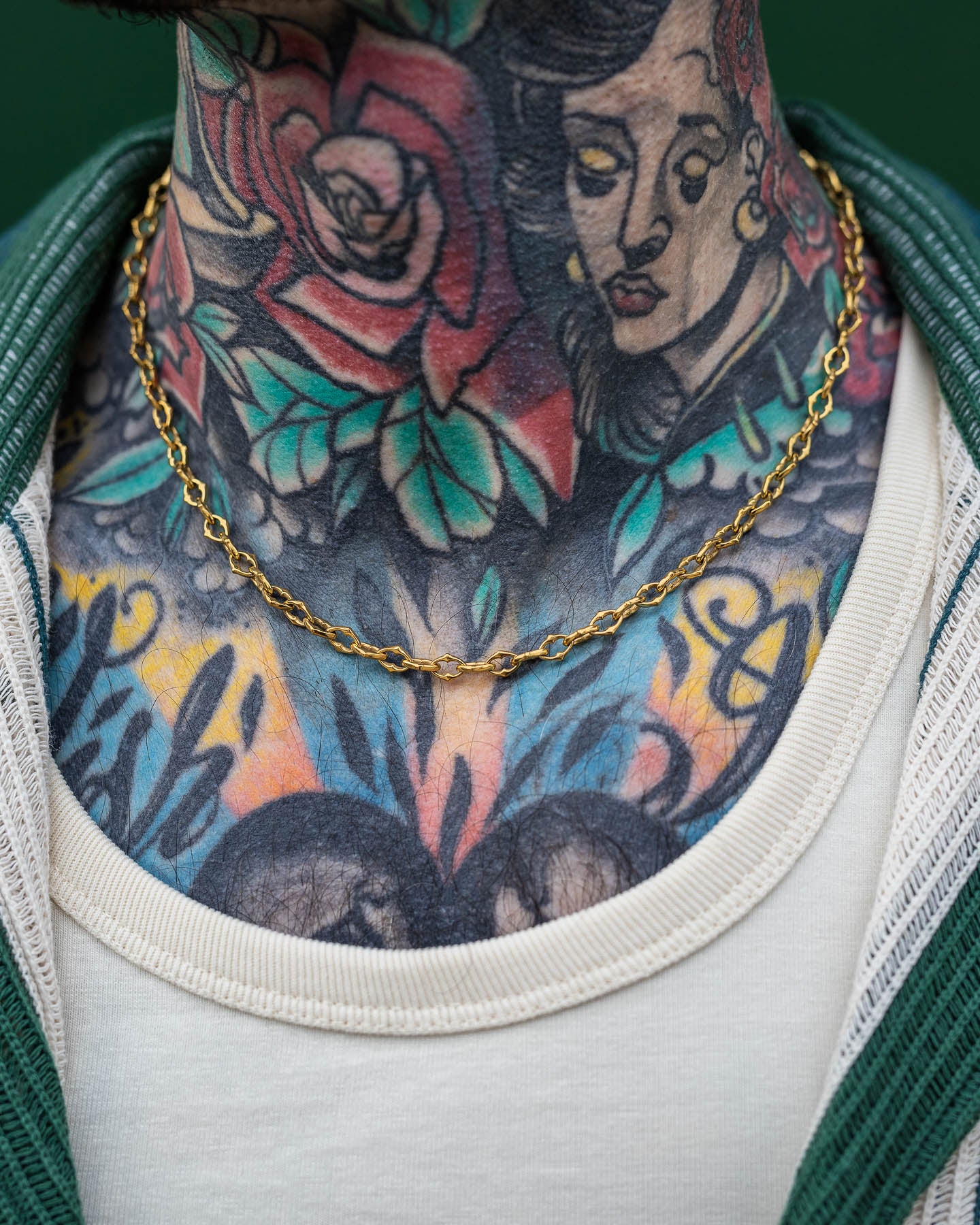 A close-up of a tattooed neck and upper chest, showcasing colorful designs and "The Cathedral" 6mm Spiked Chain (Gold) necklace, worn with a white shirt and green jacket.