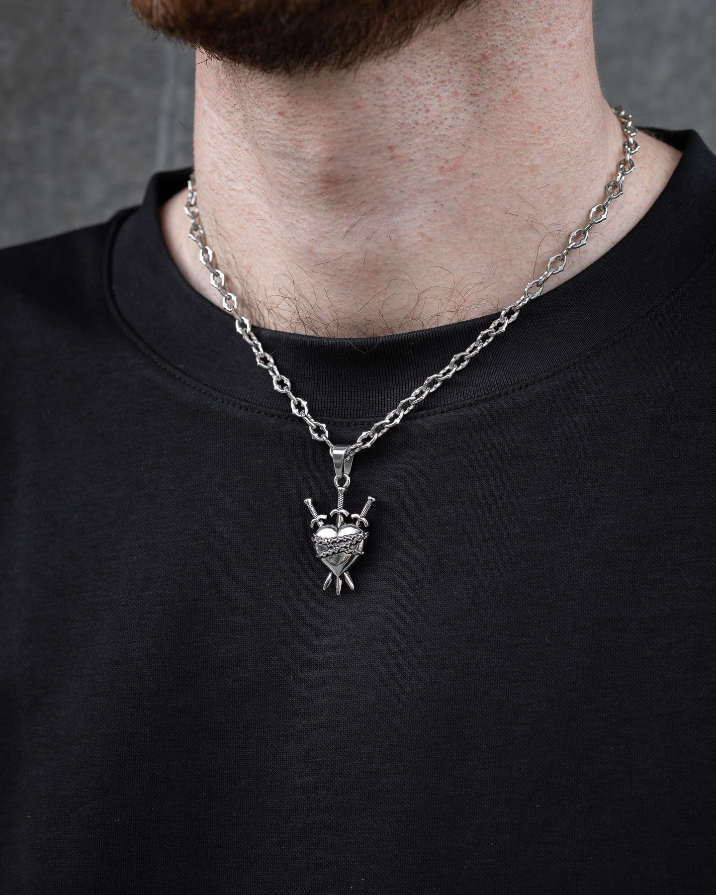 A close-up of a person wearing "The Cathedral" 6mm Spiked Chain with a heart pendant featuring two crossed swords, styled over a plain black shirt, showing their lower face and neck.