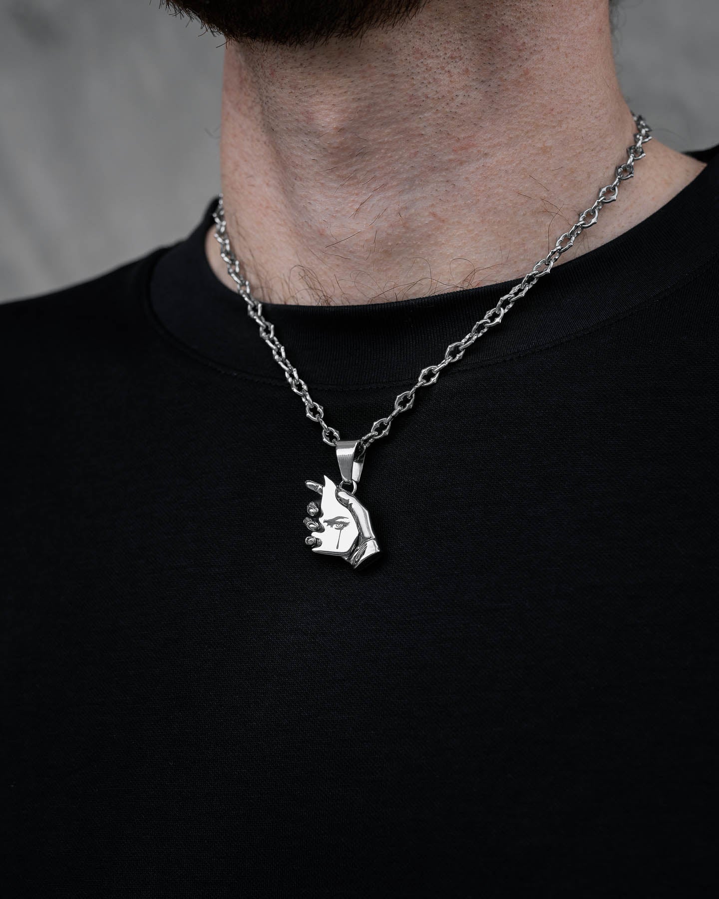 Someone wears "The Cathedral" 6mm spiked chain with a custom pendant shaped like a sculpted hand holding a small sphere, paired with a black shirt. The background is blurred gray.