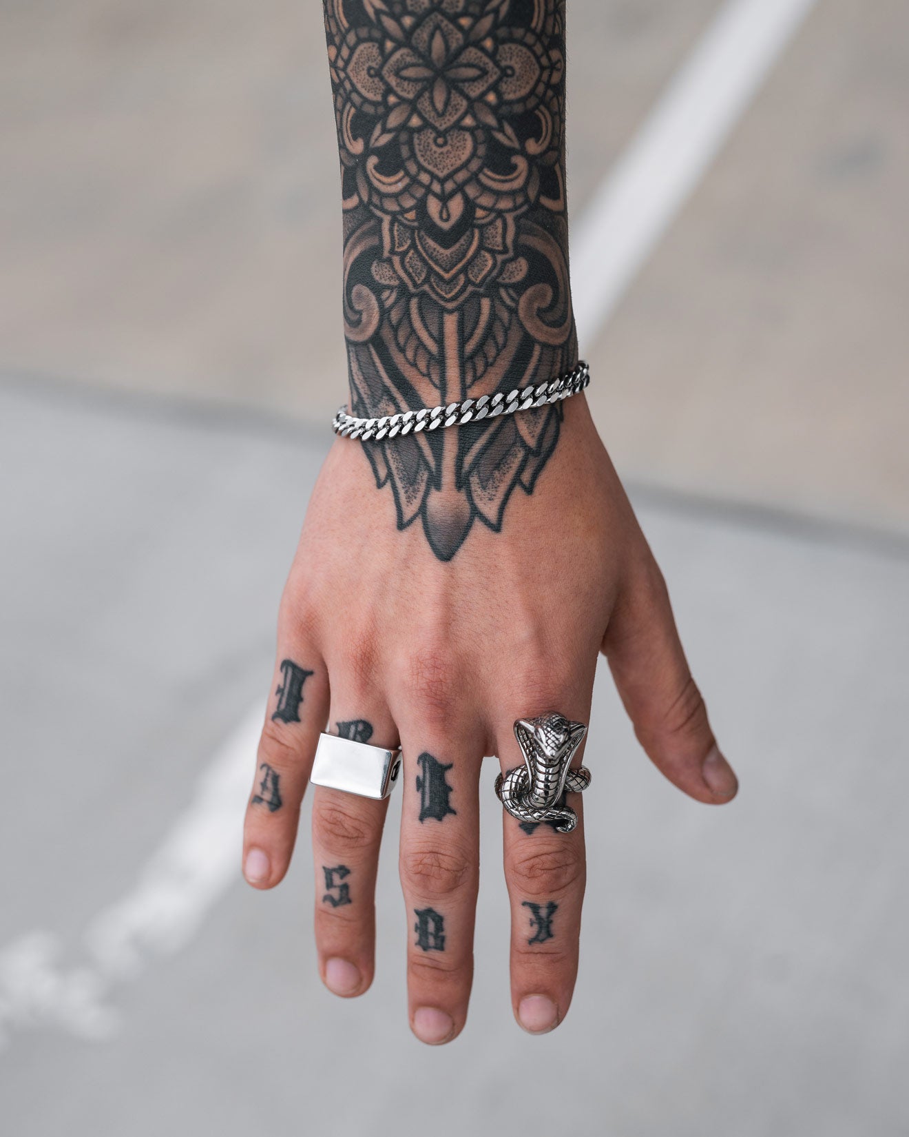 A tattooed hand with bold letter tattoos on the knuckles wears a 6mm Cuban Bracelet and two rings—one plain, one animal-head shaped. Intricate designs cover the wrist and fingers.