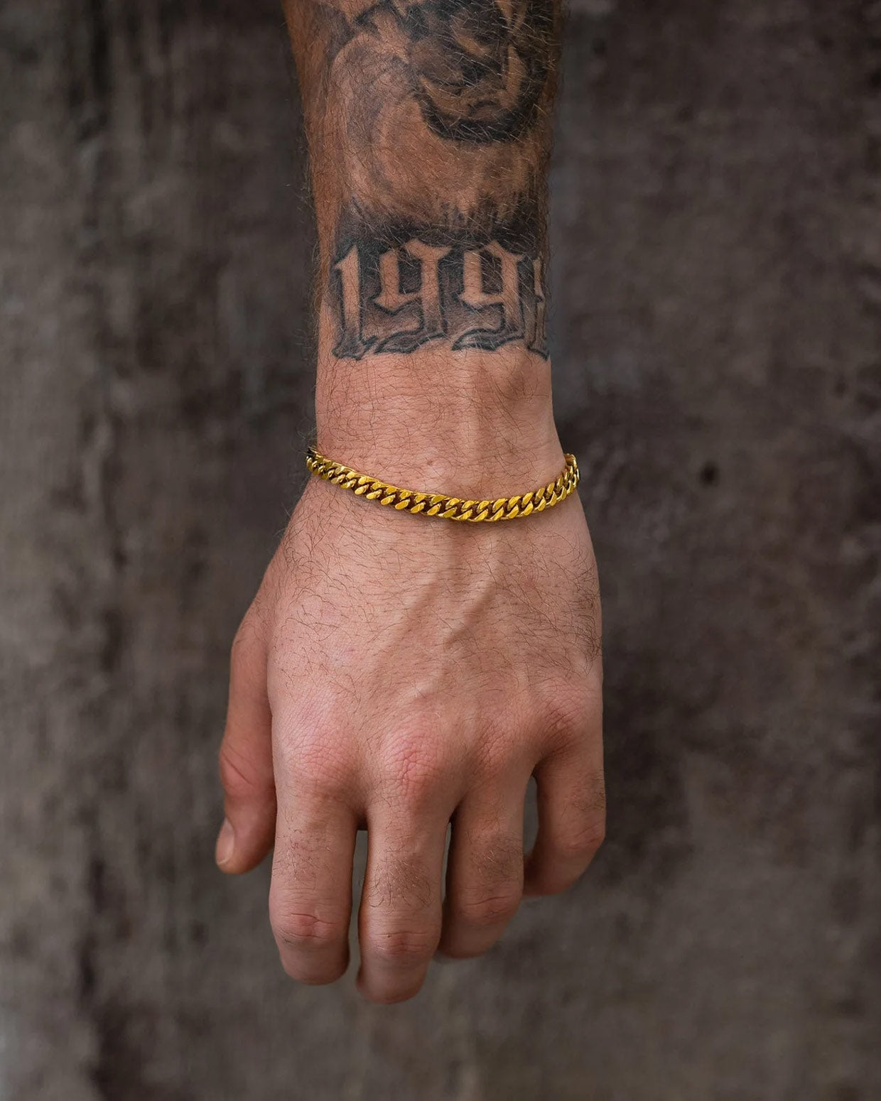 A close-up of a hand wearing the Statement Collective 6mm Cuban Bracelet (Gold), its bold 18k shine standing out next to tattoos marked "1997" and another above, all set against a dark, textured background.