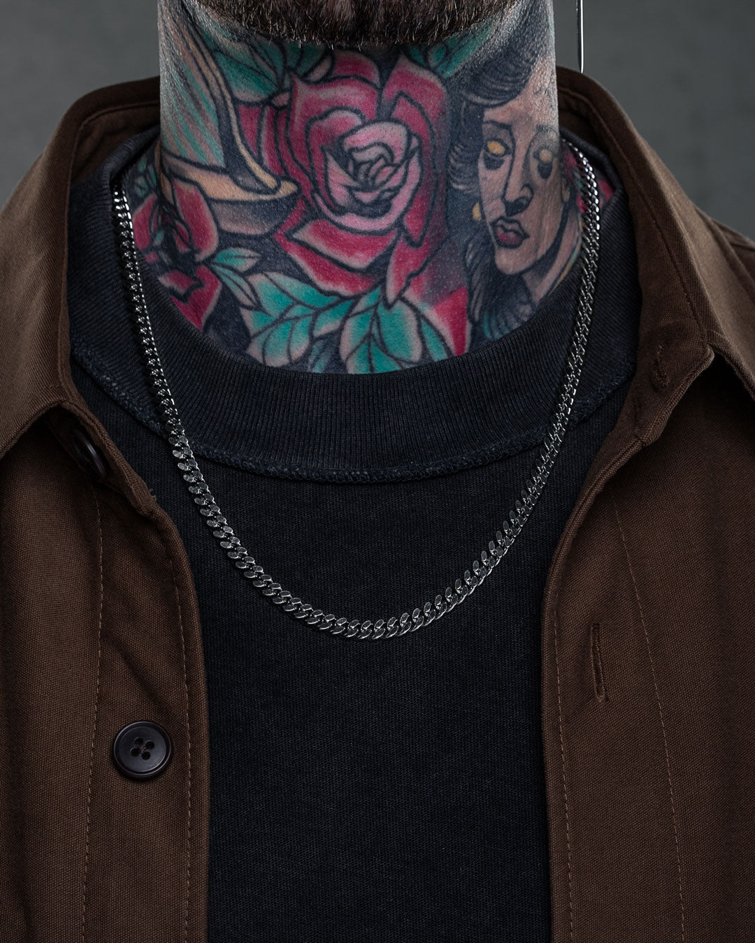 A person in a black shirt, brown jacket, and 6mm Cuban Chain shows off colorful neck tattoos featuring a rose and a woman's face.