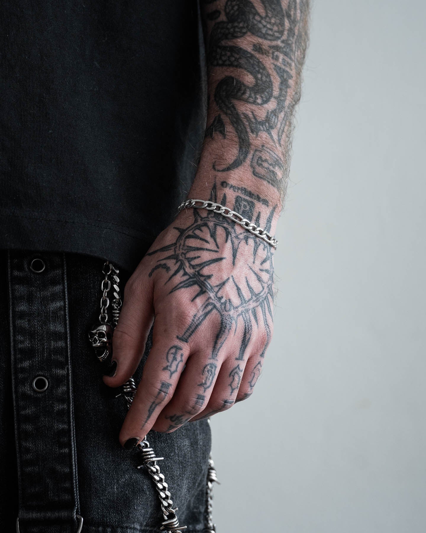 A tattooed hand with black nail polish wears a 6mm Figaro Bracelet, resting against black denim pants with a chain detail. Tattoos cover the hand, fingers, and forearm. The person completes the look with a black shirt.