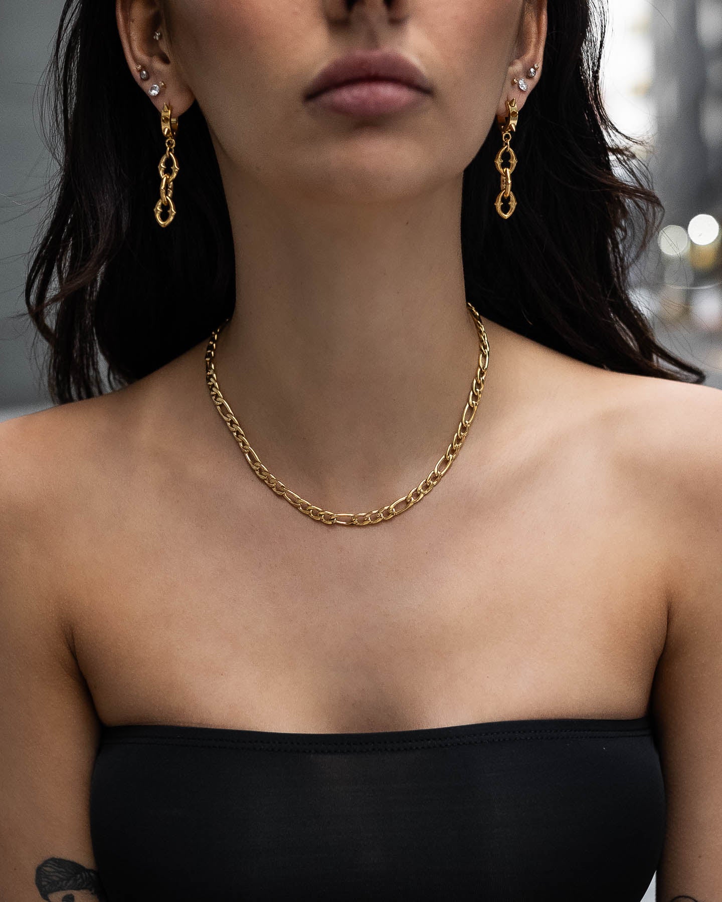 A woman in a black strapless top models the 6mm Figaro Chain (Gold) necklace and matching 18k gold-plated drop earrings. Multiple ear piercings and a blurred background keep the focus on her jewelry and neckline.