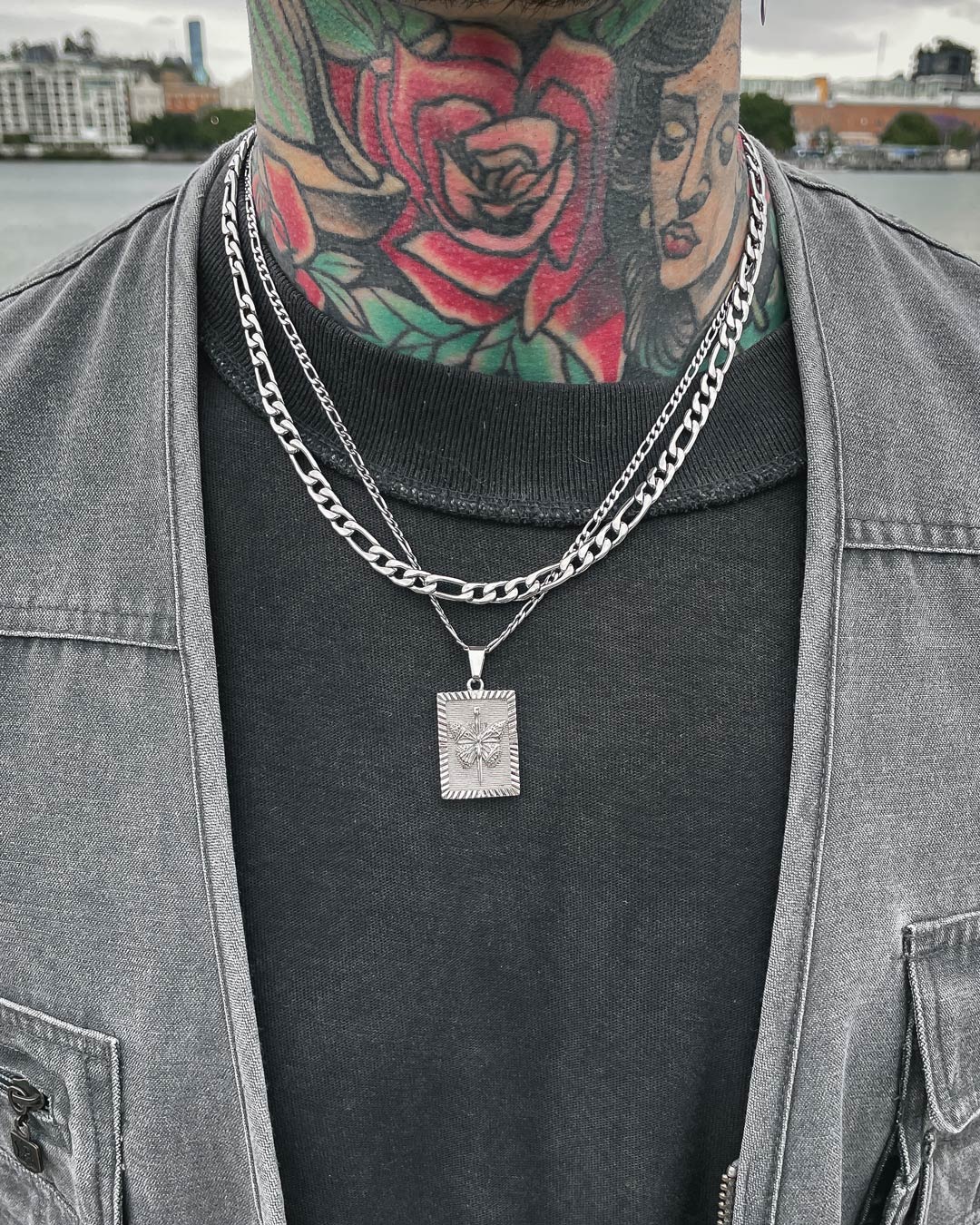 A person with colorful neck tattoos wears two silver chains, including the 6mm Figaro Chain with a square pendant, over a black shirt and grey jacket. Water and buildings are blurred in the background.