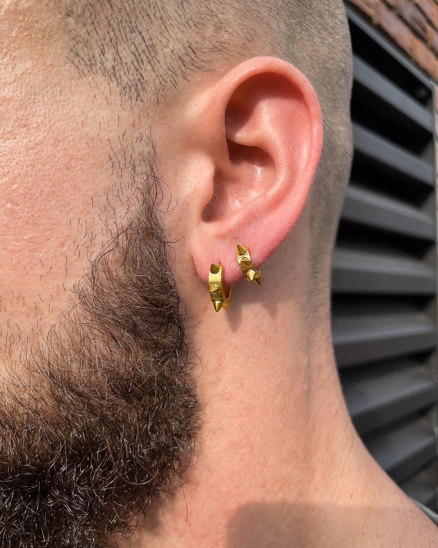 A close-up of a bearded person’s ear wearing two 6mm Spiked Hoop Earrings (Gold) in the lobe, set against a brick wall and black metal slats.