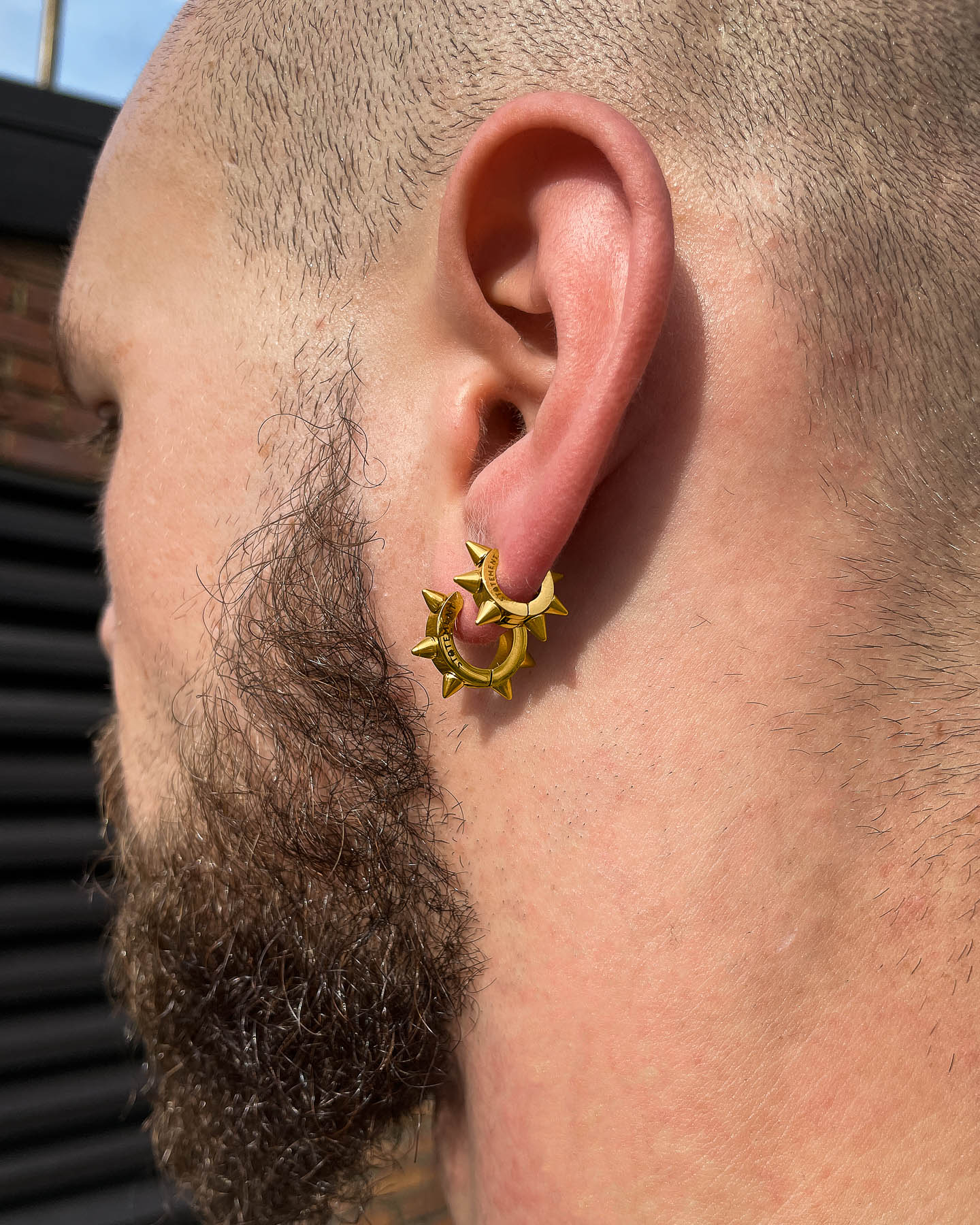 A close-up of a bearded person’s ear shows the 6mm Spiked Hoop Earrings (Gold) featuring star accents. The hypoallergenic gold hoops stand out against a brick wall and black horizontal slats.