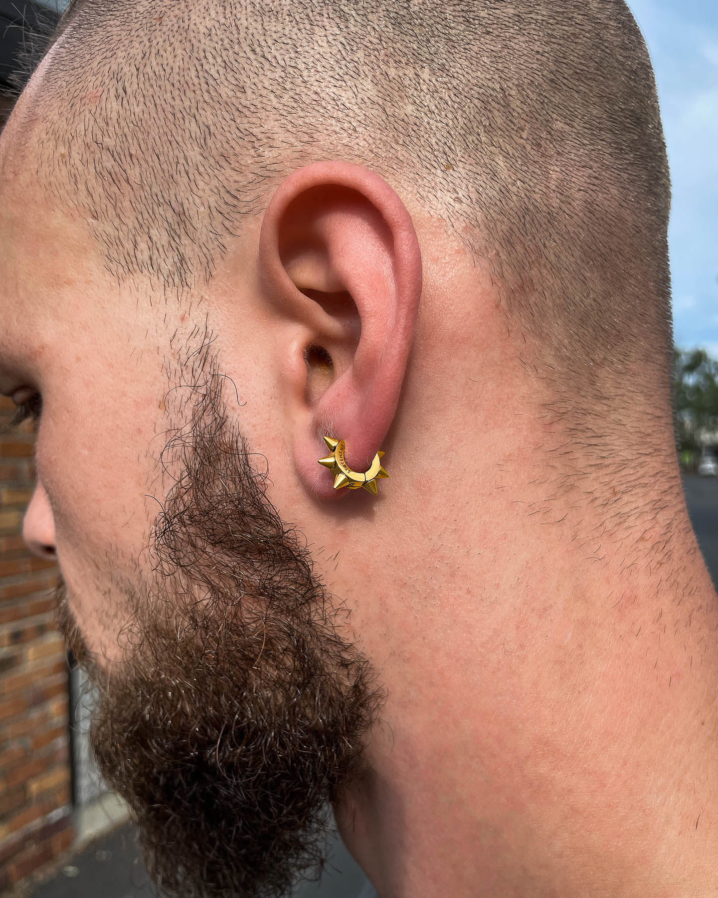 Close-up of a person's ear wearing 6mm Spiked Hoop Earrings (Gold) with a trimmed beard and short hair. Set against an outdoor brick wall and trees, these hypoallergenic hoops add an edgy touch to any look.