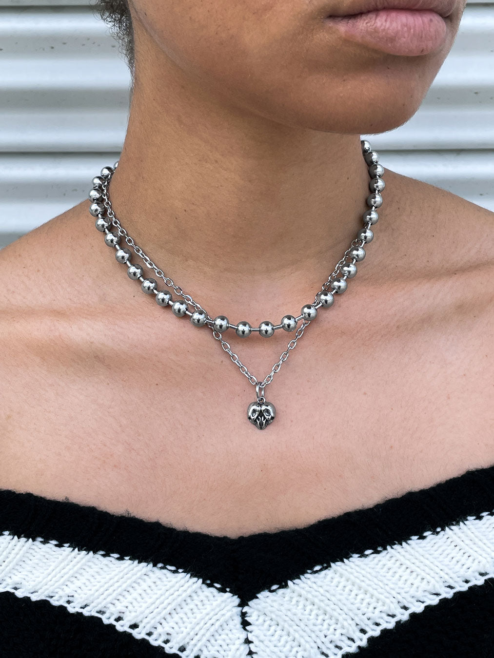 A person wearing layered silver necklaces, including a 7mm Ball Chain with large beads and a heart pendant, paired with a black and white off-the-shoulder zigzag top—classic mall goth jewelry style.