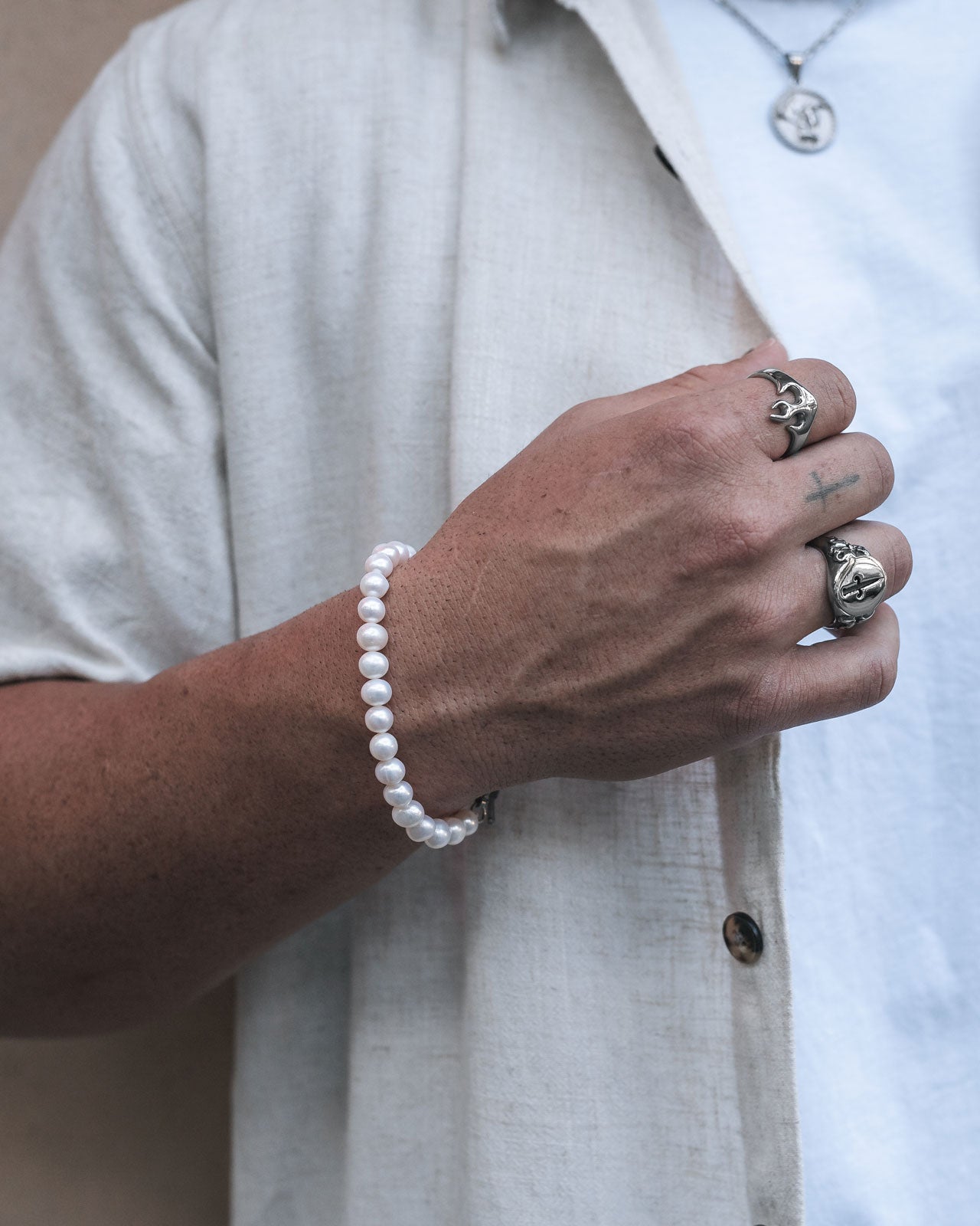 A person wearing a light short-sleeve shirt and white t-shirt shows their hand adorned with silver rings and the 8mm Freshwater Pearl Bracelet. A pendant necklace is partially visible around their neck.