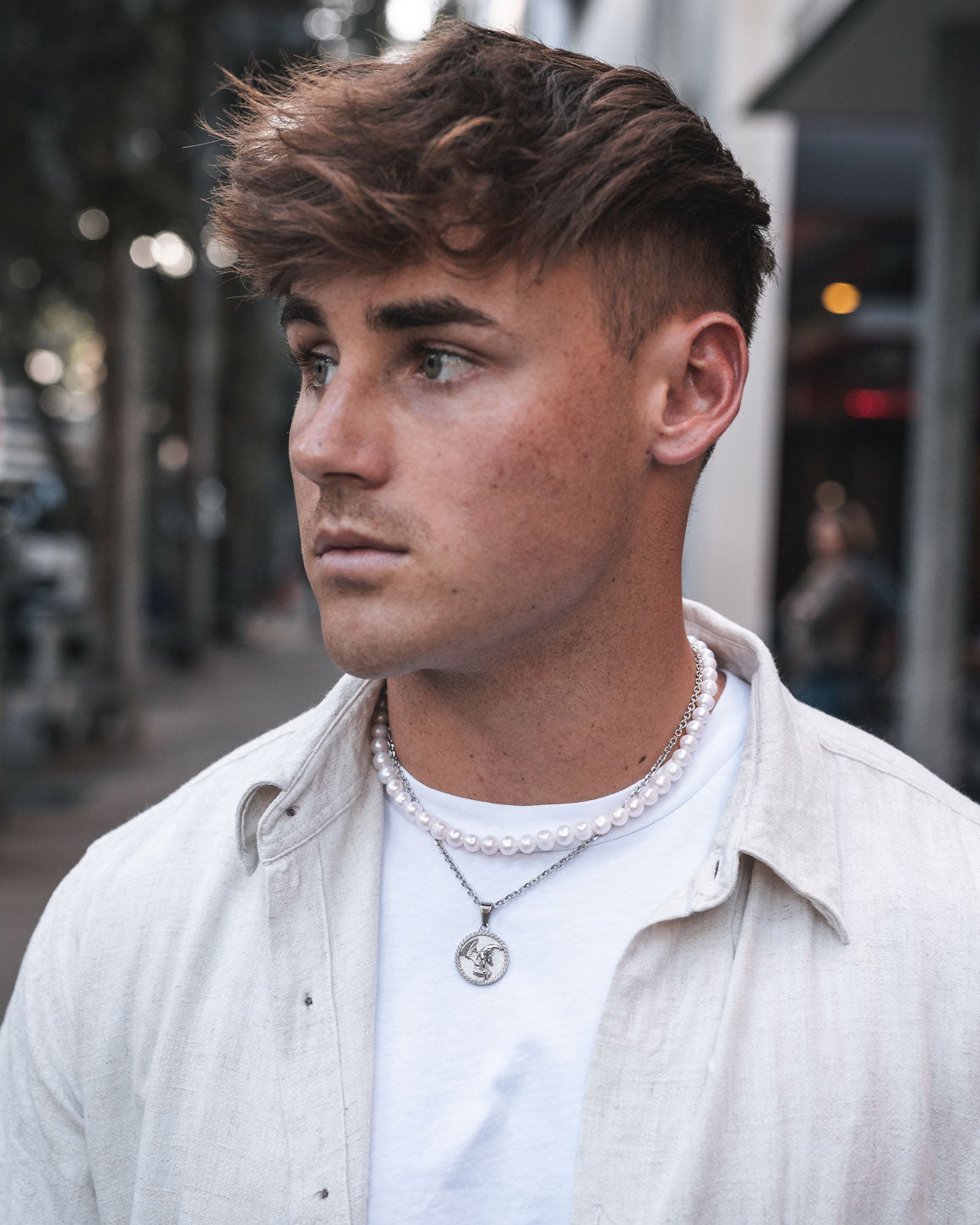 A young man with short brown hair and blue eyes stands on a city street, wearing a light shirt over a white tee and layered necklaces, including the 8mm Freshwater Pearl Necklace with a round pendant.