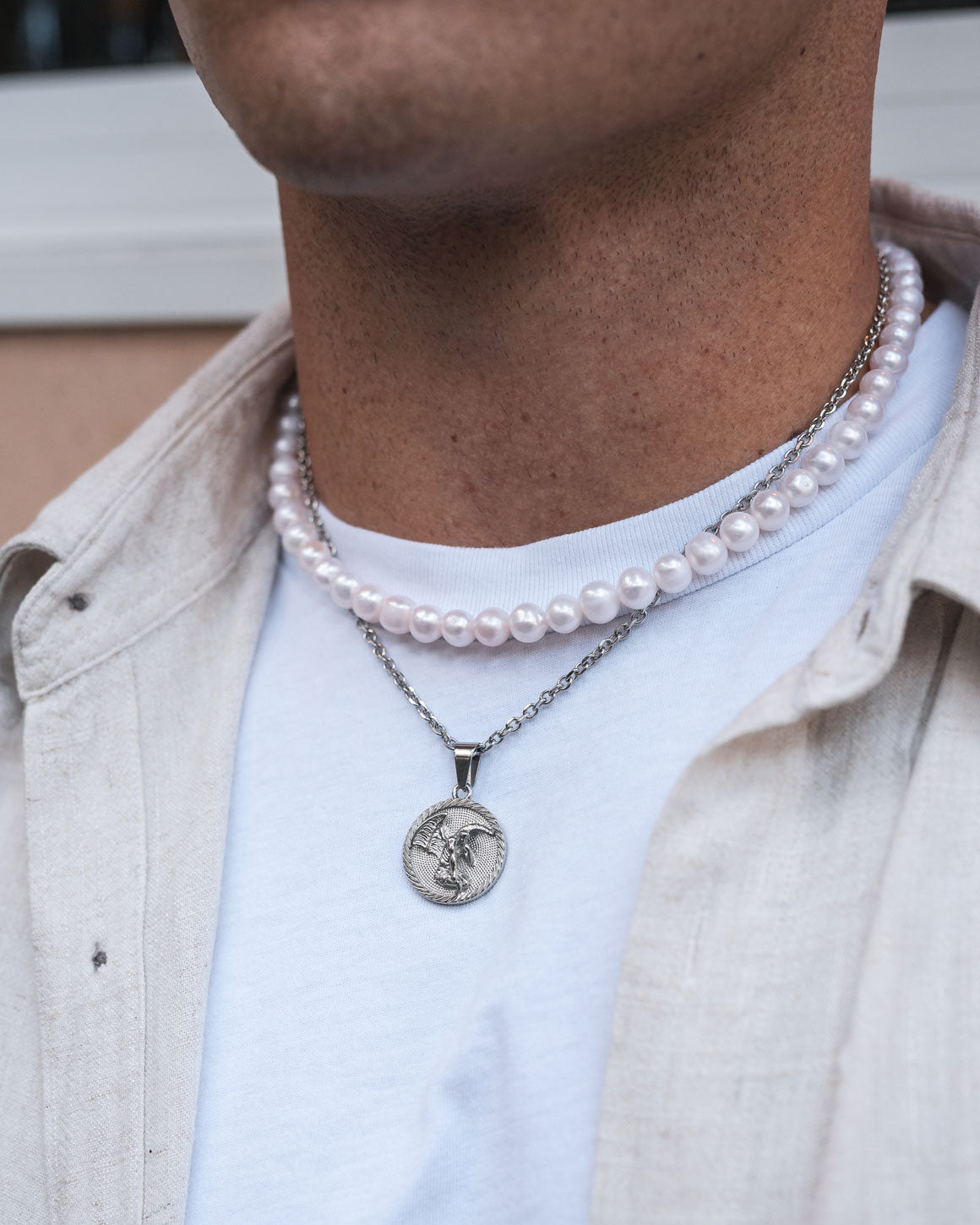 A person wearing a white shirt and cream overshirt is shown layering an 8mm Freshwater Pearl Necklace with a silver chain necklace featuring a round pendant. The focus is on the neck and the pearl jewelry.