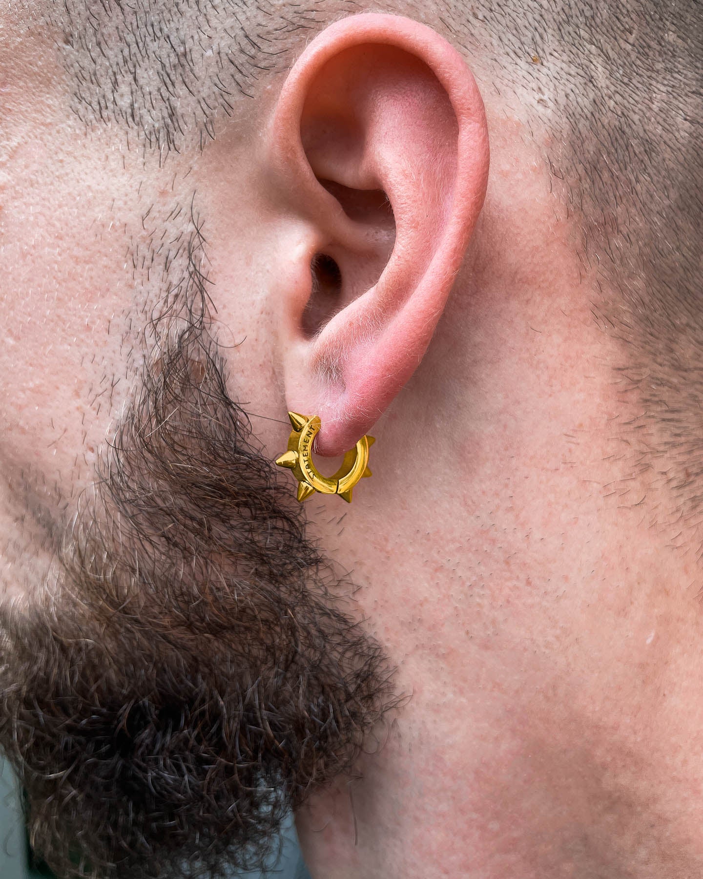 A close-up of a person's ear with 8mm Spiked Hoop Earrings (Gold). With a short beard and trimmed hair, they effortlessly showcase this subtle yet edgy accessory.