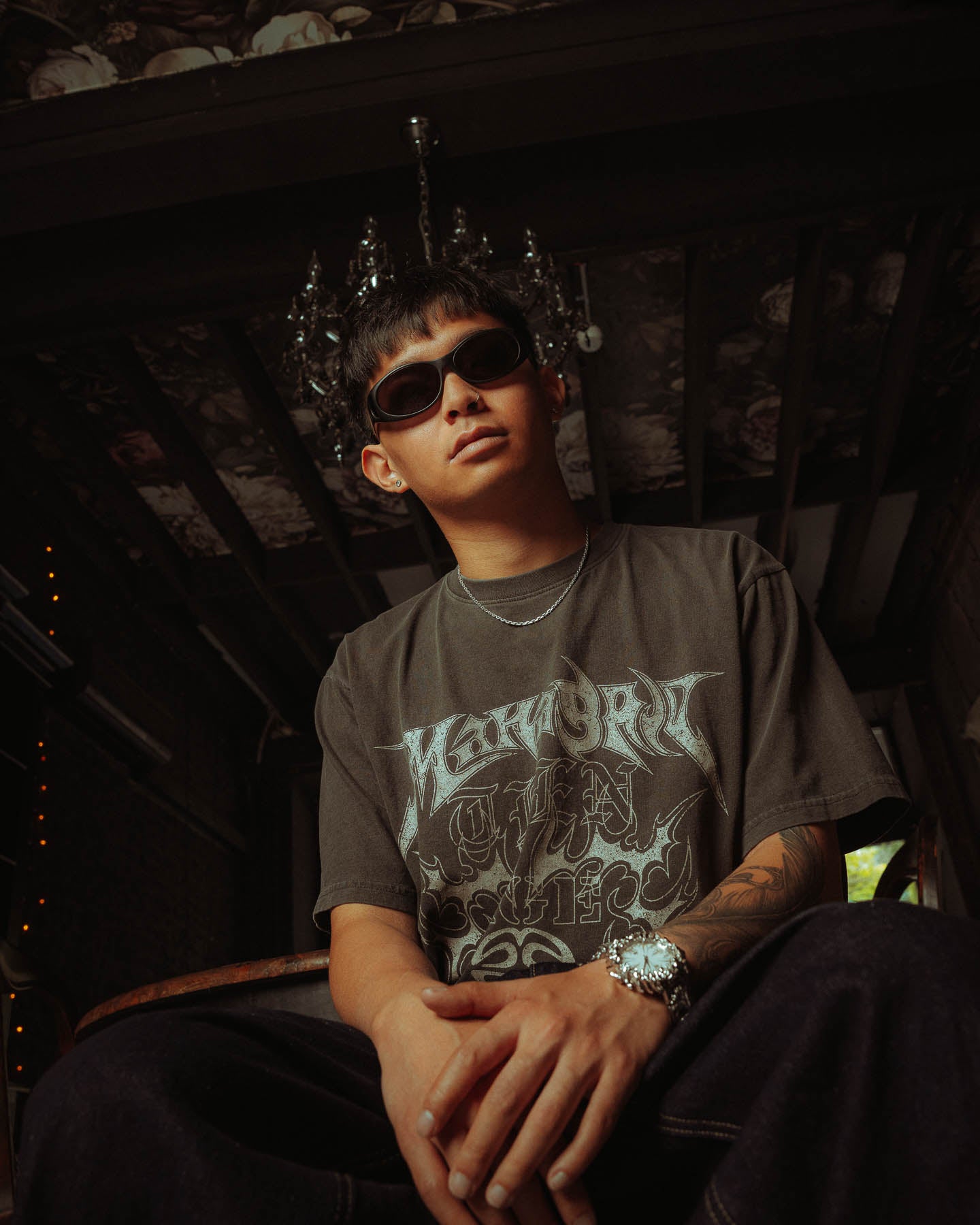 A person wearing dark sunglasses, a graphic t-shirt, a watch, and a 925 Sterling Silver 3mm Cable Chain necklace sits indoors under a chandelier, confidently looking at the camera with hands clasped in a moody, artistic setting.