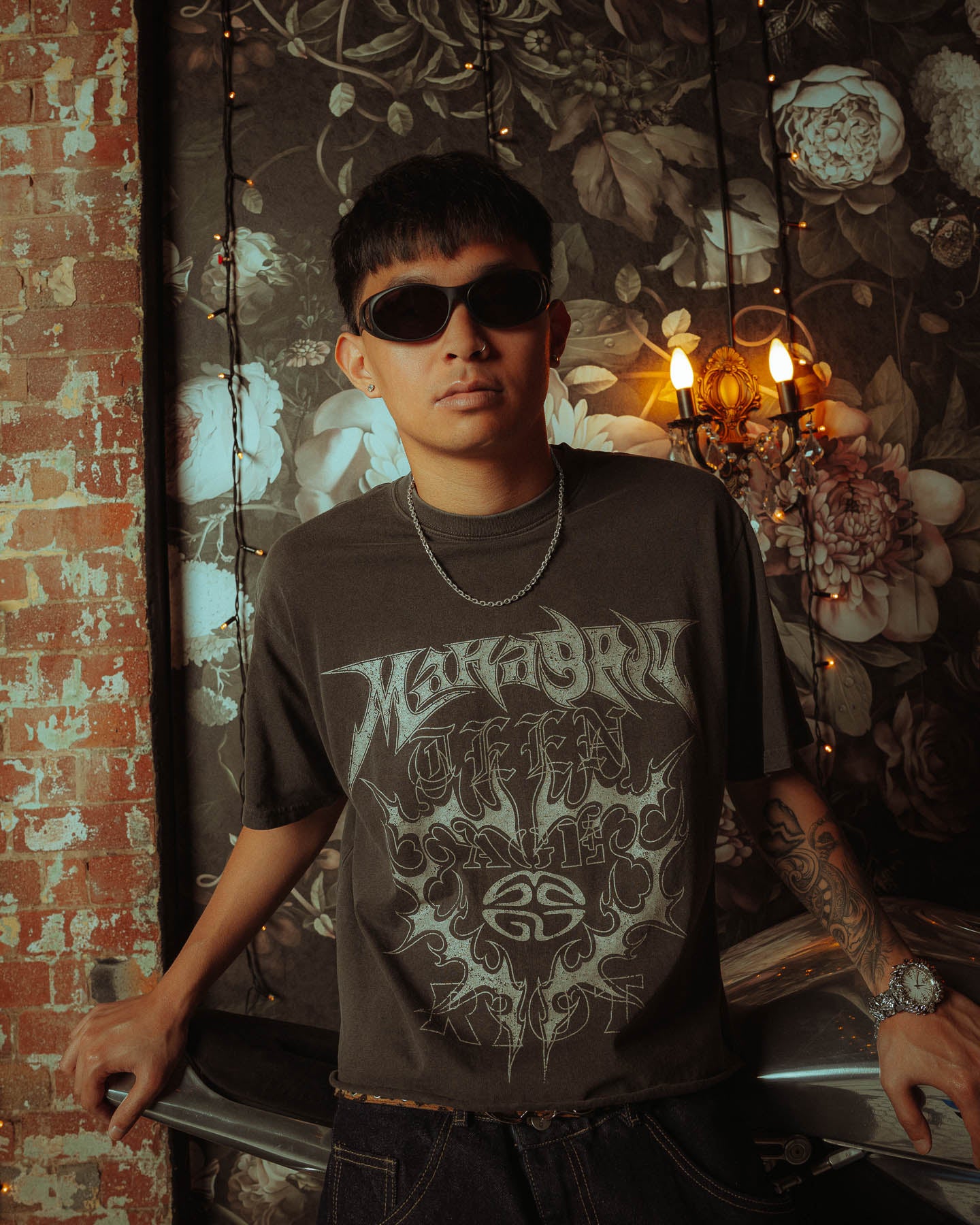 Wearing dark sunglasses, a graphic tee, and a 925 Sterling Silver 4.5mm Cable Chain, a person leans on a silver surface with floral wallpaper, string lights, a sconce, and exposed brick in the background.