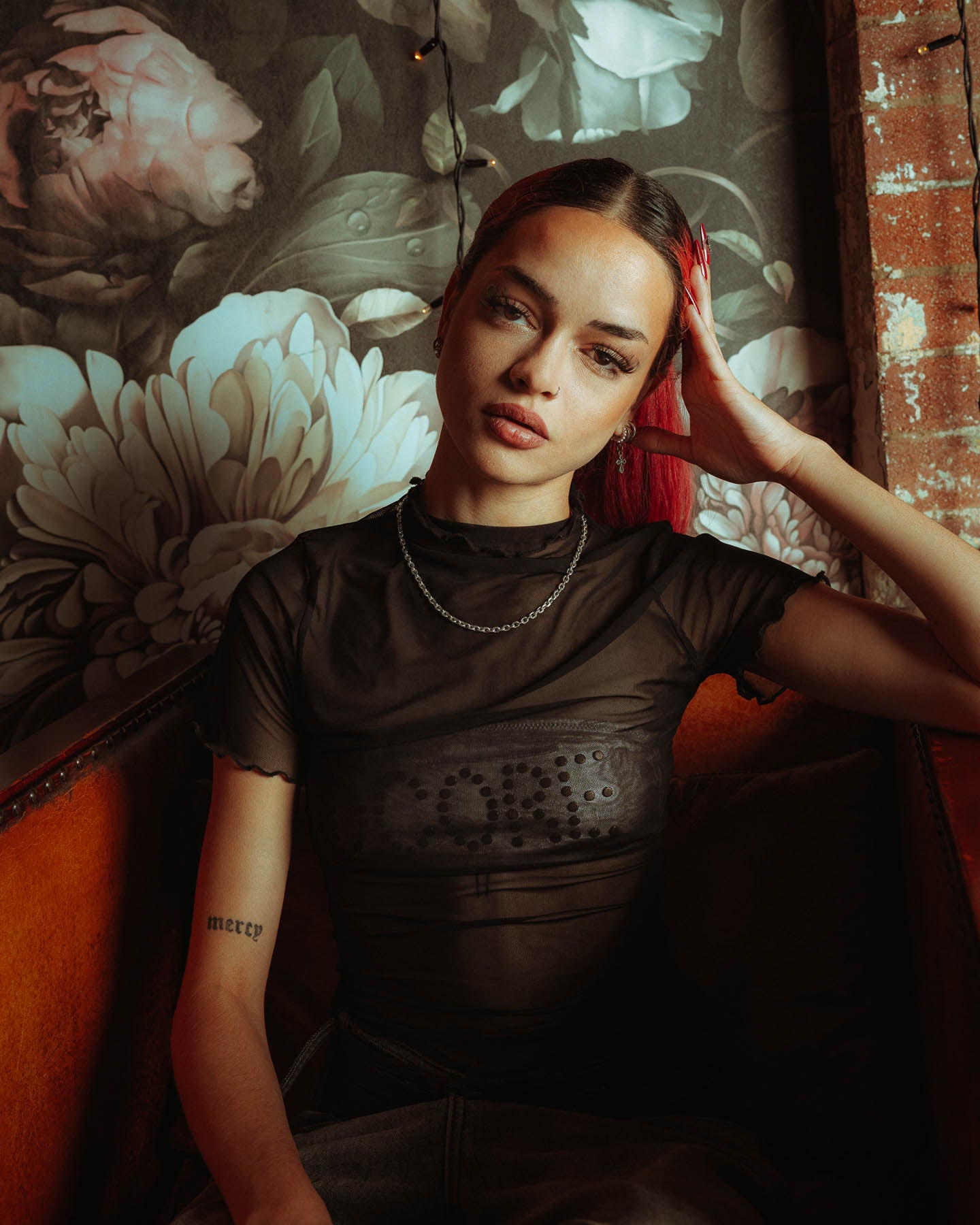 A person with straight red hair sits on a couch, one hand on their head, wearing a sheer black top and a 925 Sterling Silver 4.5mm Cable Chain necklace. Floral wallpaper and exposed brick are in the background.