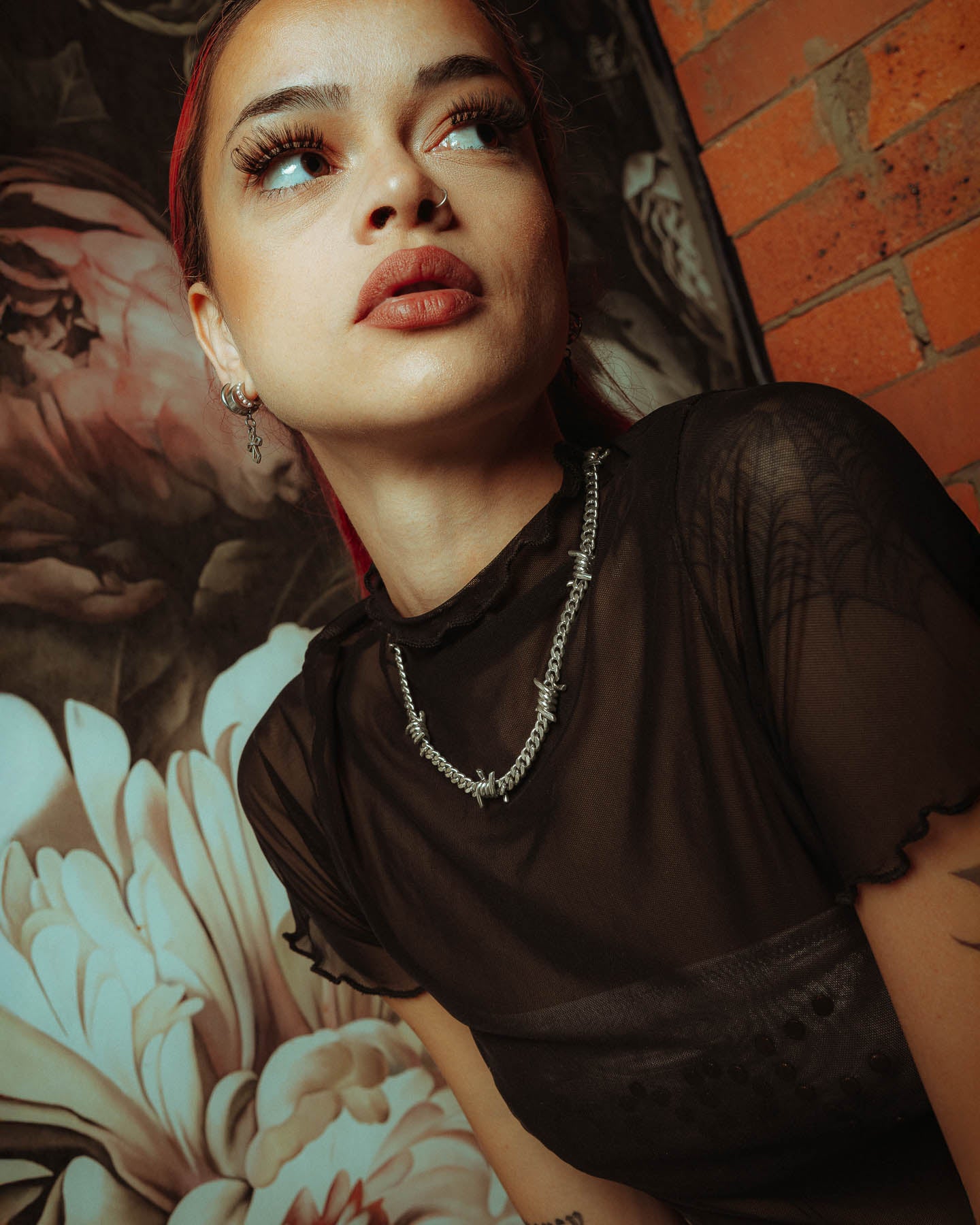 A woman with red hair and bold makeup wears a sheer black top and the 925 Sterling Silver 6mm Barbed Wire Cuban Chain necklace, sitting thoughtfully before a floral mural and brick wall.