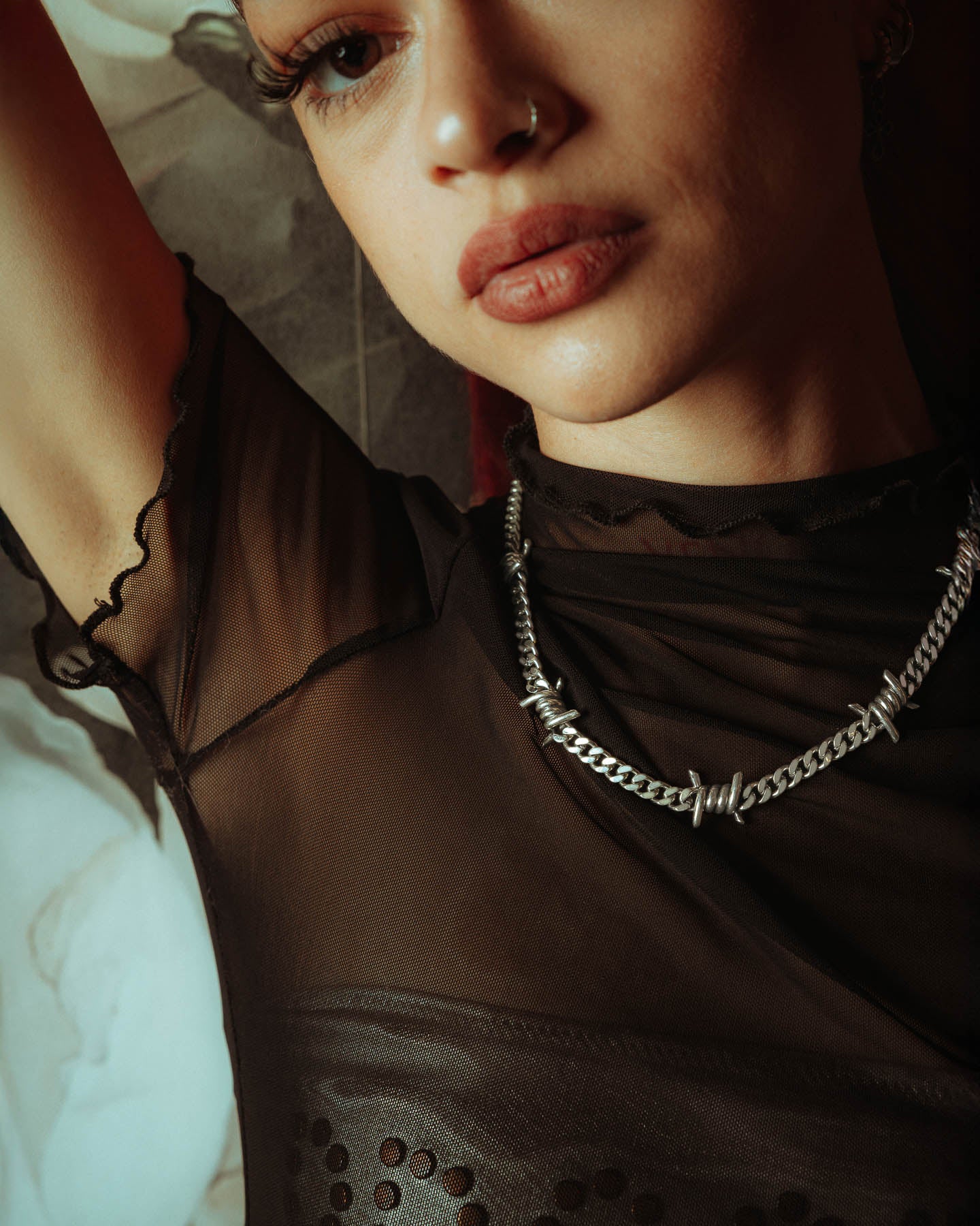 A person in a sheer black top poses with one arm raised, showing soft makeup, a nose ring, and an edgy look featuring the 925 Sterling Silver 6mm Barbed Wire Cuban Chain.