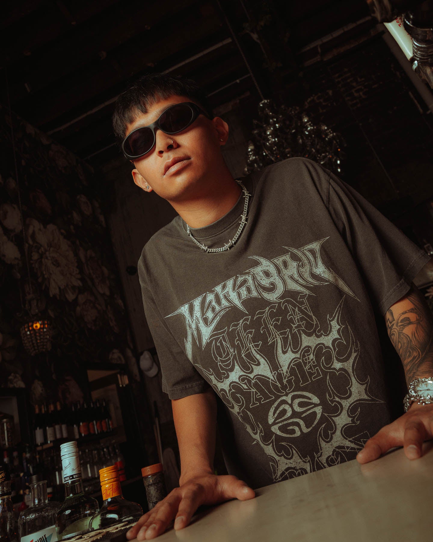 A young man in dark sunglasses and a black graphic tee leans on a bar counter, sporting an alternative vibe with a 925 Sterling Silver 6mm Barbed Wire Cuban Chain. Bottles and dim lighting in the background create a moody atmosphere.