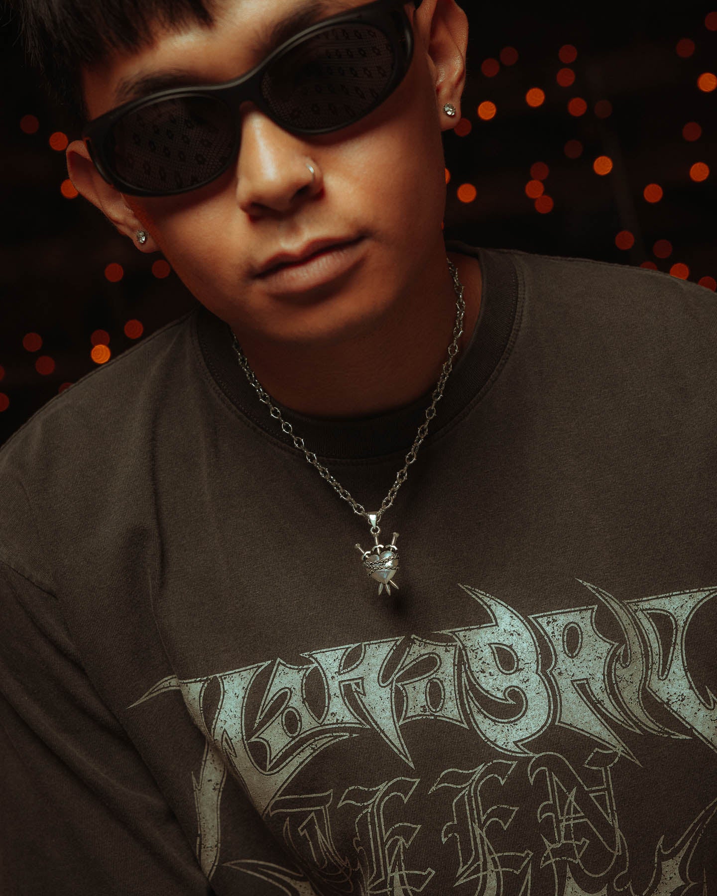 A person in dark sunglasses and a black graphic tee wears the 925 Sterling Silver "The Cathedral" 6mm Spiked Chain with a custom pendant, standing before warm, blurry lights.