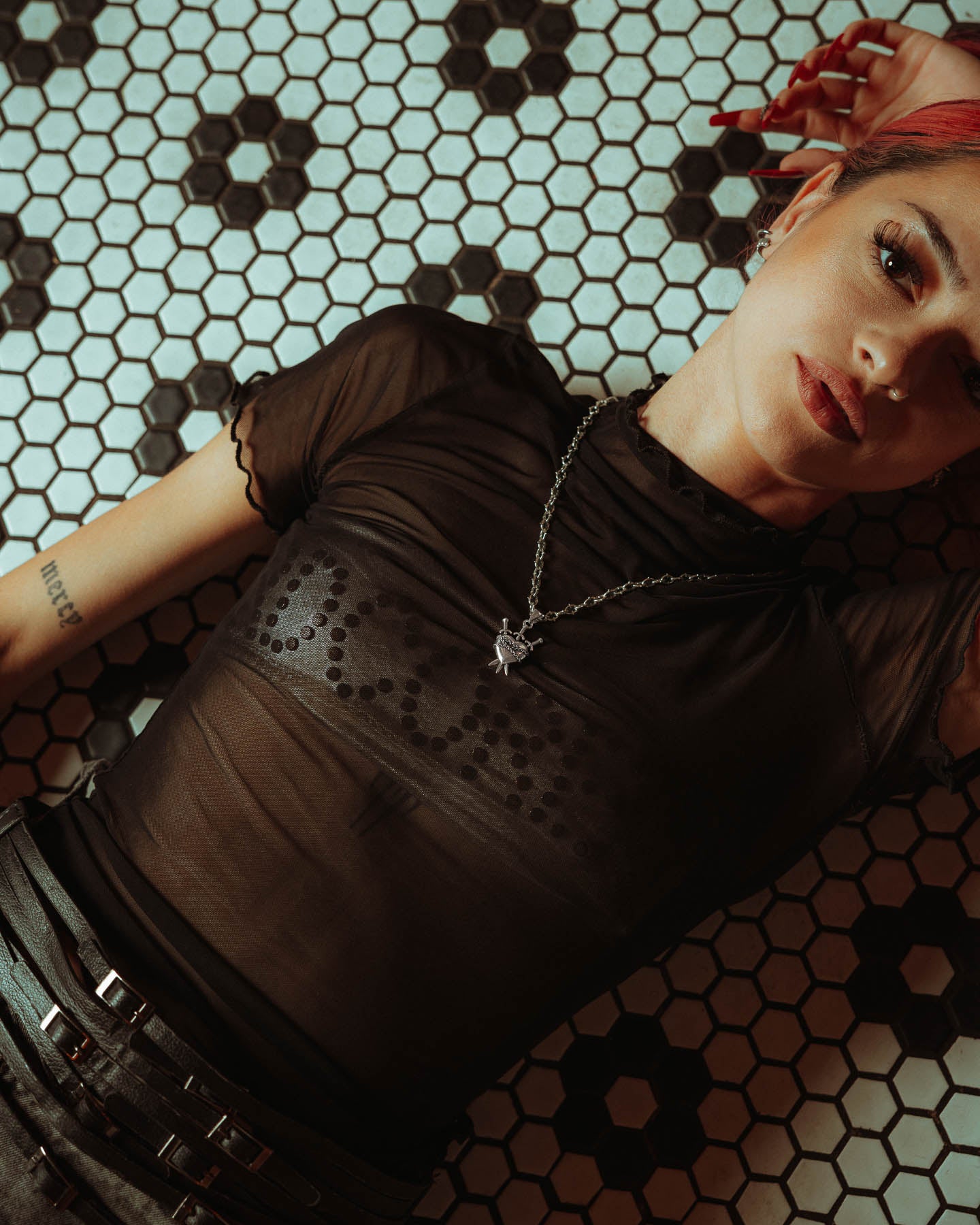 A person with pink hair and dark eye makeup in a sheer black top lies on a black-and-white floral hex tile floor, gazing at the camera. They wear layered necklaces, including the 925 Sterling Silver "The Cathedral" 6mm Spiked Chain with custom pendant.