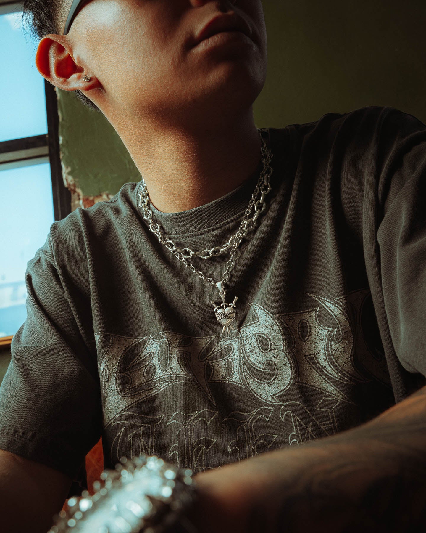 A person wearing a dark graphic tee, glasses, and the 925 Sterling Silver "The Cathedral" 6mm Spiked Chain with their choice of custom pendant sits by a window. Dramatic lighting casts bold shadows over their face and shirt.