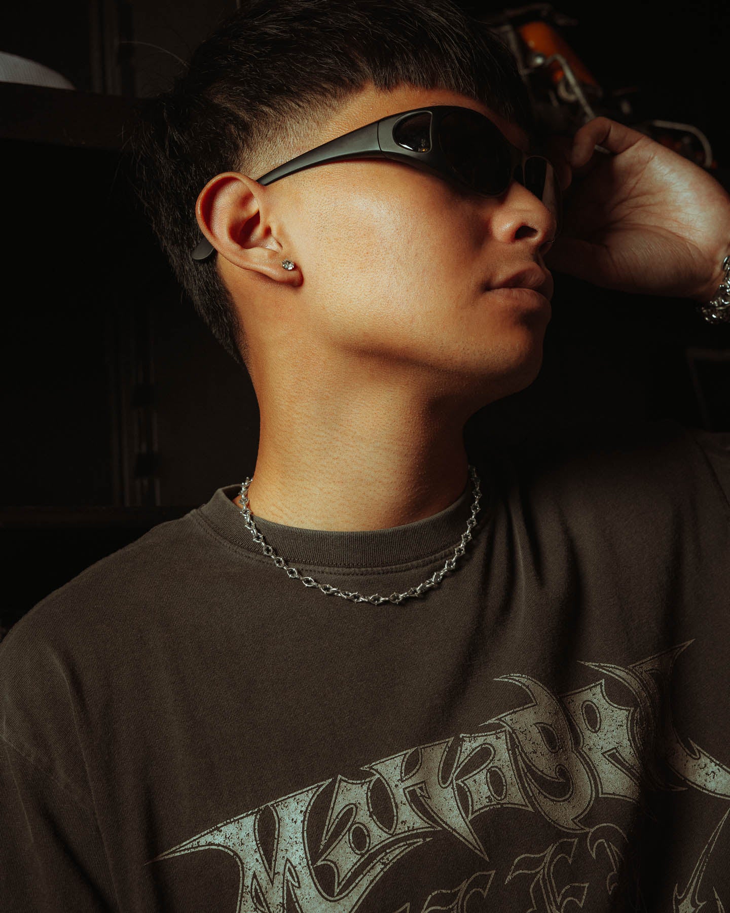 A person wearing dark sunglasses, a graphic t-shirt, an earring, and "The Cathedral" 6mm Spiked 925 Sterling Silver Chain looks to the side while adjusting their glasses against a dark background.