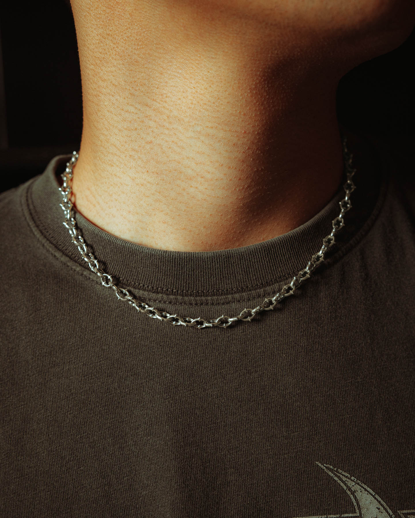 A close-up of a person’s neck wearing a dark t-shirt and the 925 Sterling Silver "The Cathedral" 6mm Spiked Chain. Warm lighting highlights the chain’s elegant shine, the shirt's fabric, and the skin’s texture.