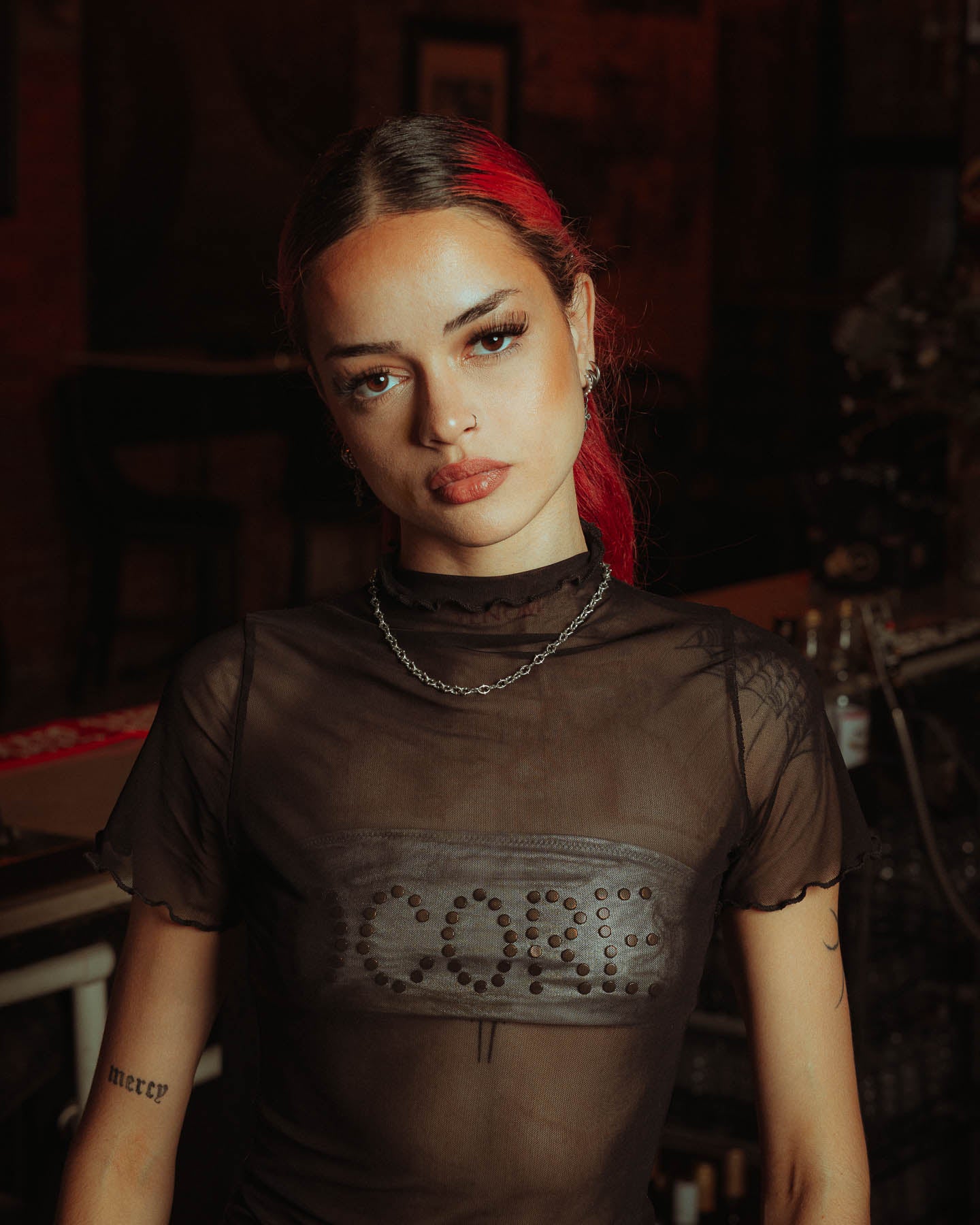 A person with long red and dark hair, wearing a sheer black top and "The Cathedral" 6mm Spiked Chain in 925 Sterling Silver, stands indoors in dim lighting. Their expression is neutral, showcasing tattoos on their arms.