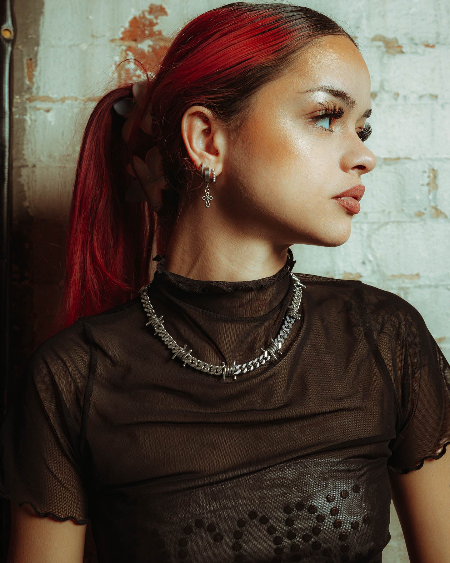 A woman with long red hair in a ponytail wears a sheer black top and layered statement jewellery, including the 925 Sterling Silver 9mm Barbed Wire Cuban Chain and a cross earring. She poses in profile against a white brick wall, looking to the right.