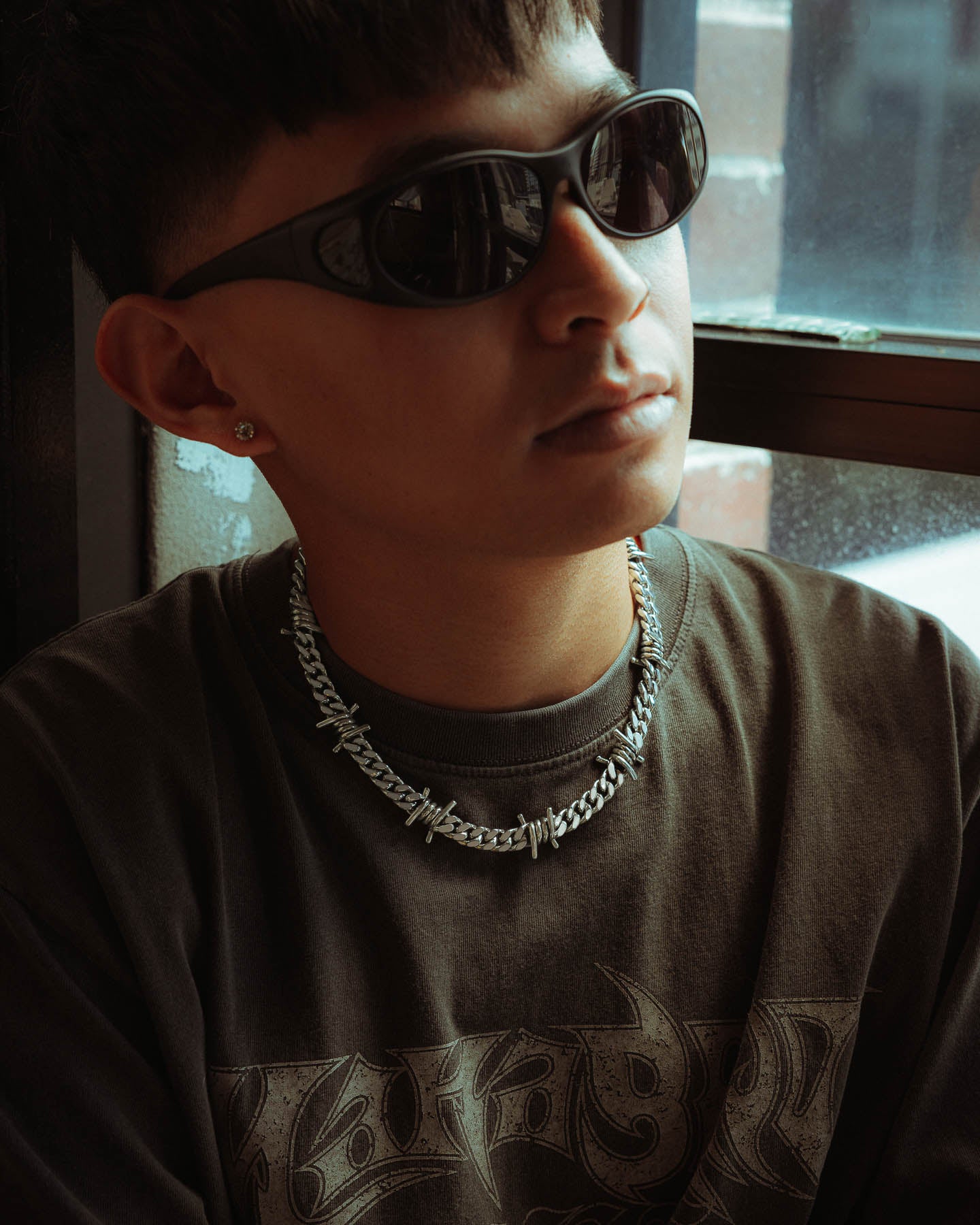 A young person in dark sunglasses, a graphic black t-shirt, and a 925 Sterling Silver 9mm Barbed Wire Cuban Chain sits by a sunlit window, their stud earring gleaming—showcasing true statement jewellery.