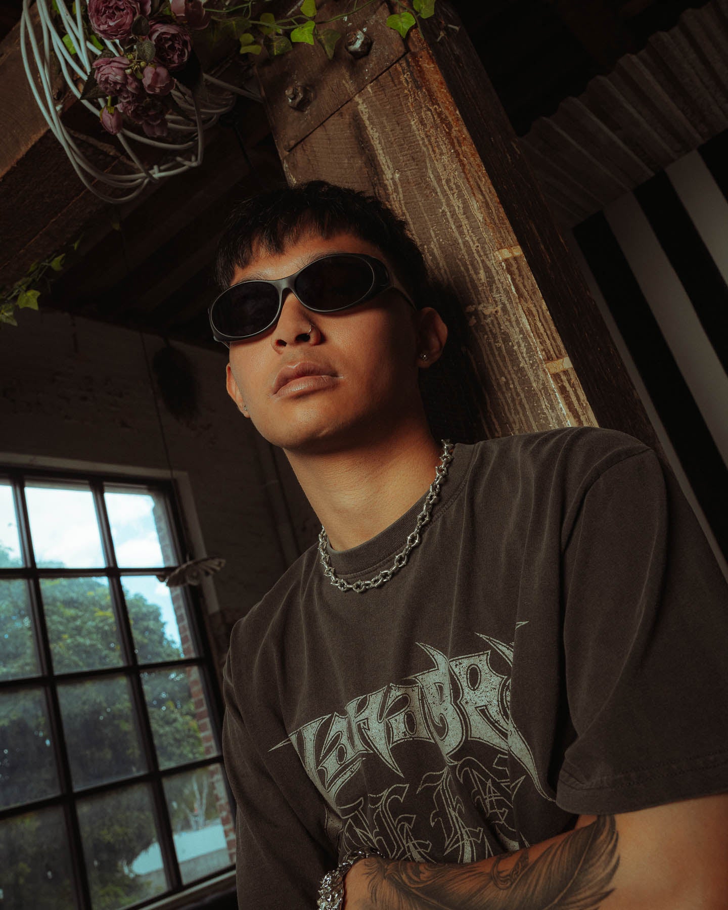 A young man in dark sunglasses and a graphic tee leans against a wooden beam indoors, showing off the 925 Sterling Silver "The Cathedral" 9mm Spiked Chain necklace. He has short dark hair, a forearm tattoo, and greenery is visible through the window behind him.