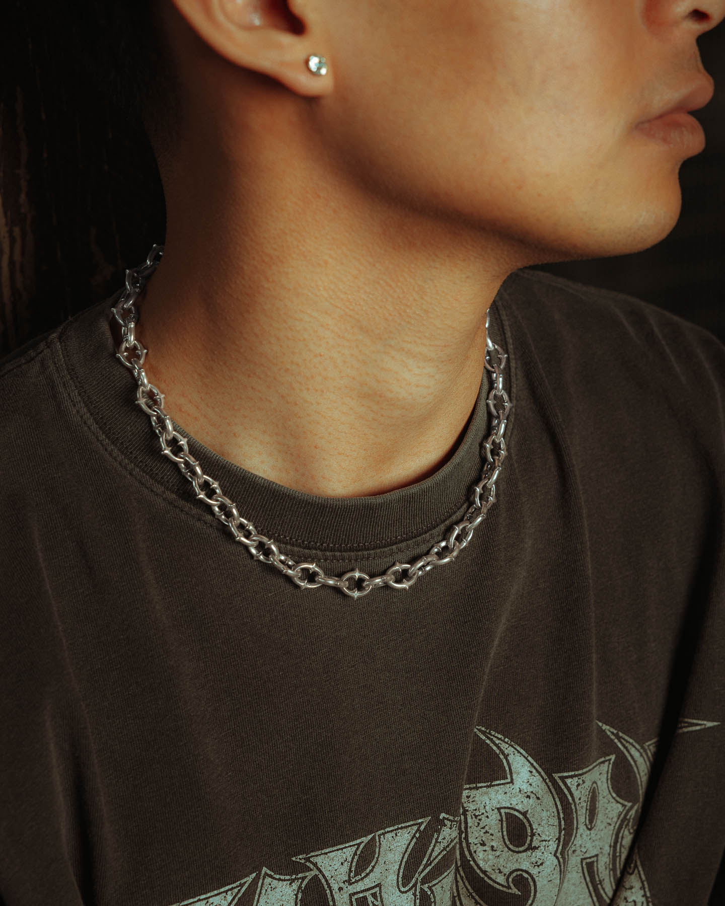 A person wears a dark graphic T-shirt and the 925 Sterling Silver "The Cathedral" 9mm Spiked Chain. A small stud earring is also visible. The photo is cropped to show only the lower half of their face and neck.
